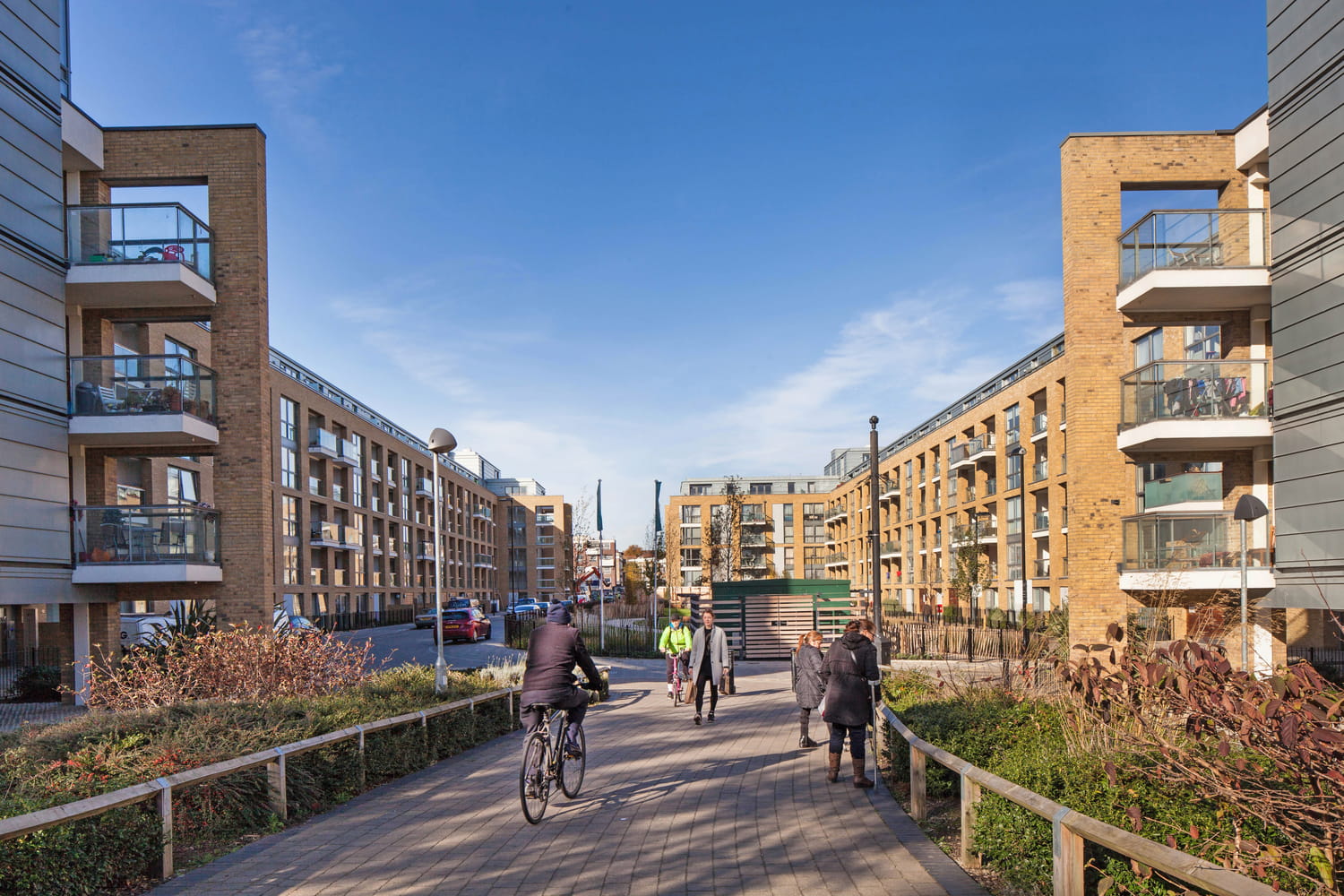 Modern residential buildings with balconies lining a pedestrian pathway, featuring people walking and cycling under a clear blue sky.