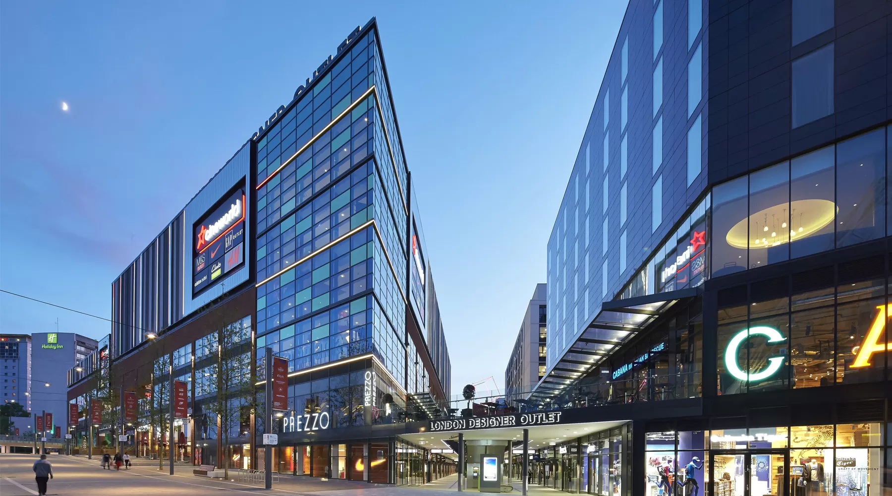 Modern retail complex featuring glass facades, illuminated signage, and storefronts, with a clear evening sky.