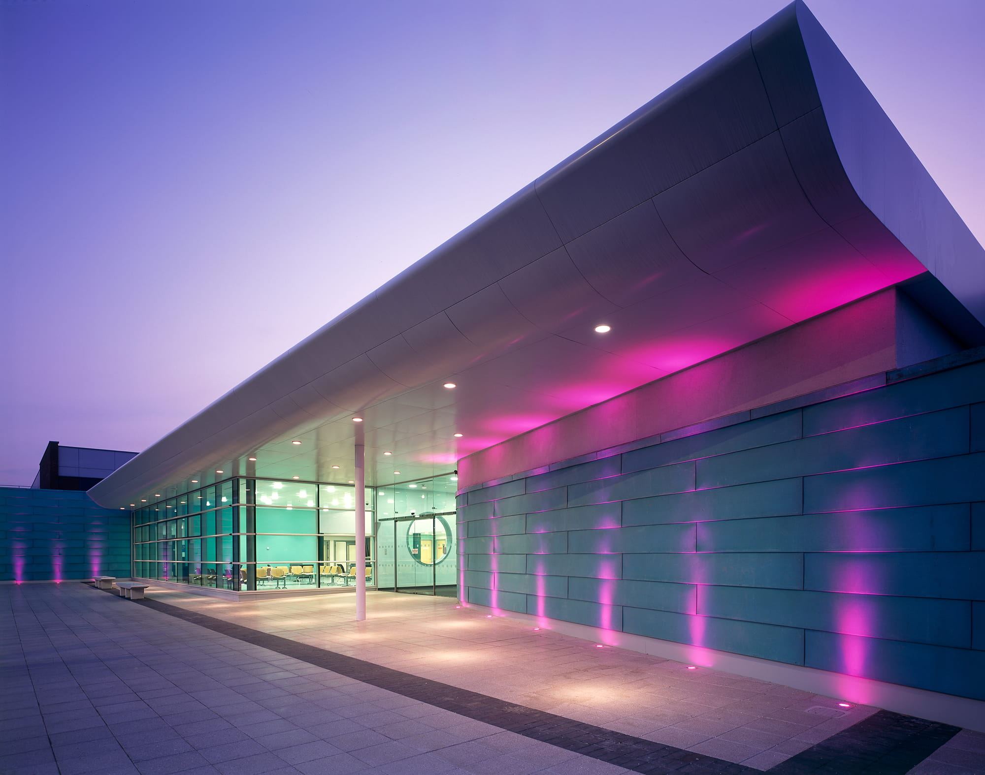 Modern healthcare facility entrance with glass walls and purple lighting at dusk.