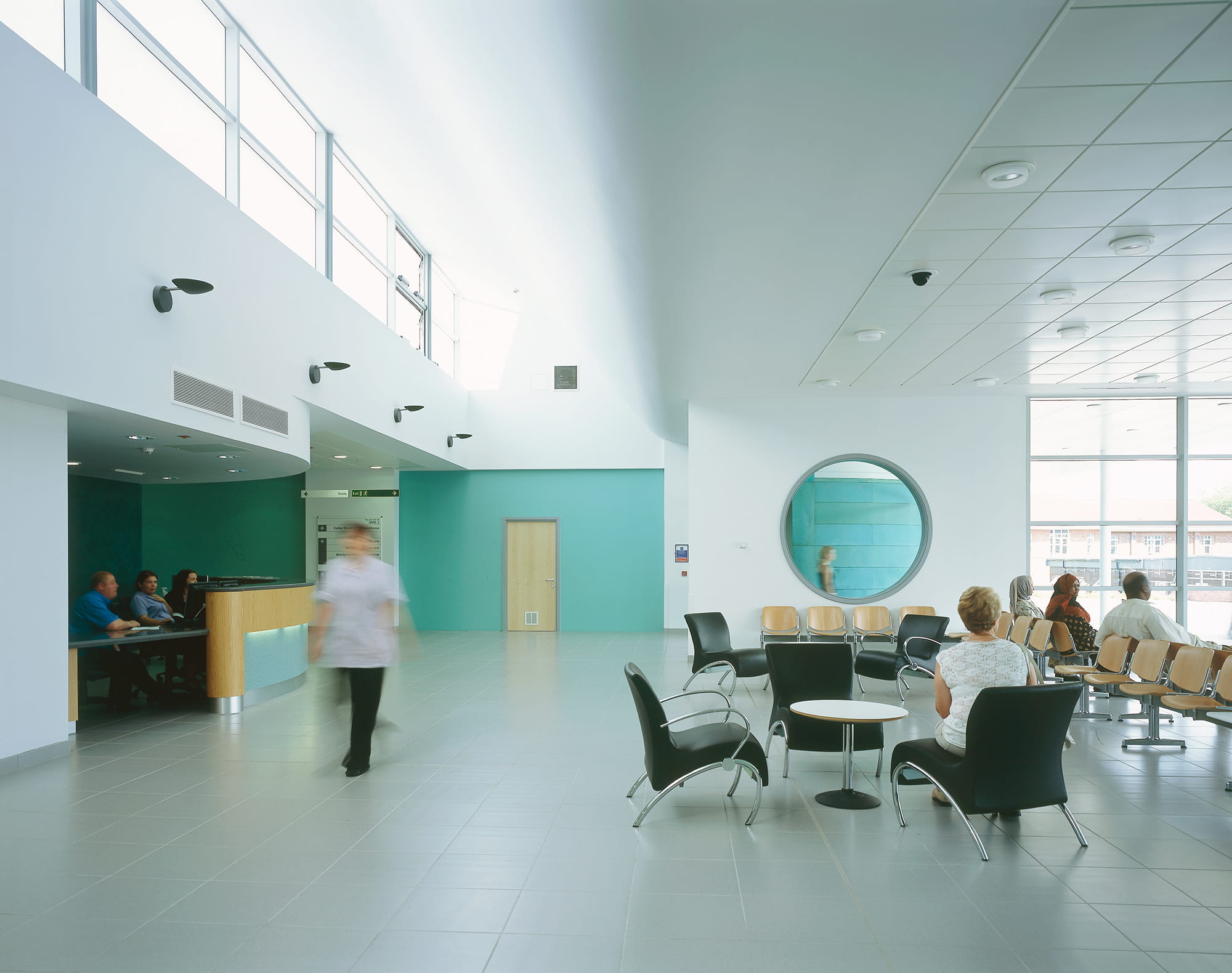 Brightly lit healthcare reception area with a curved desk, seating for patients, and a large circular mirror on the wall.