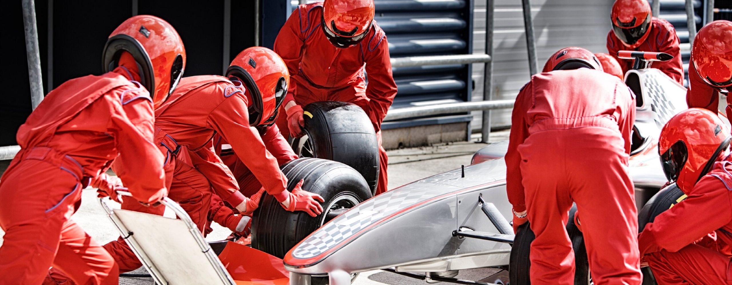 A team of mechanics in red uniforms and helmets is changing tires on a Formula 1 car in a pit stop.