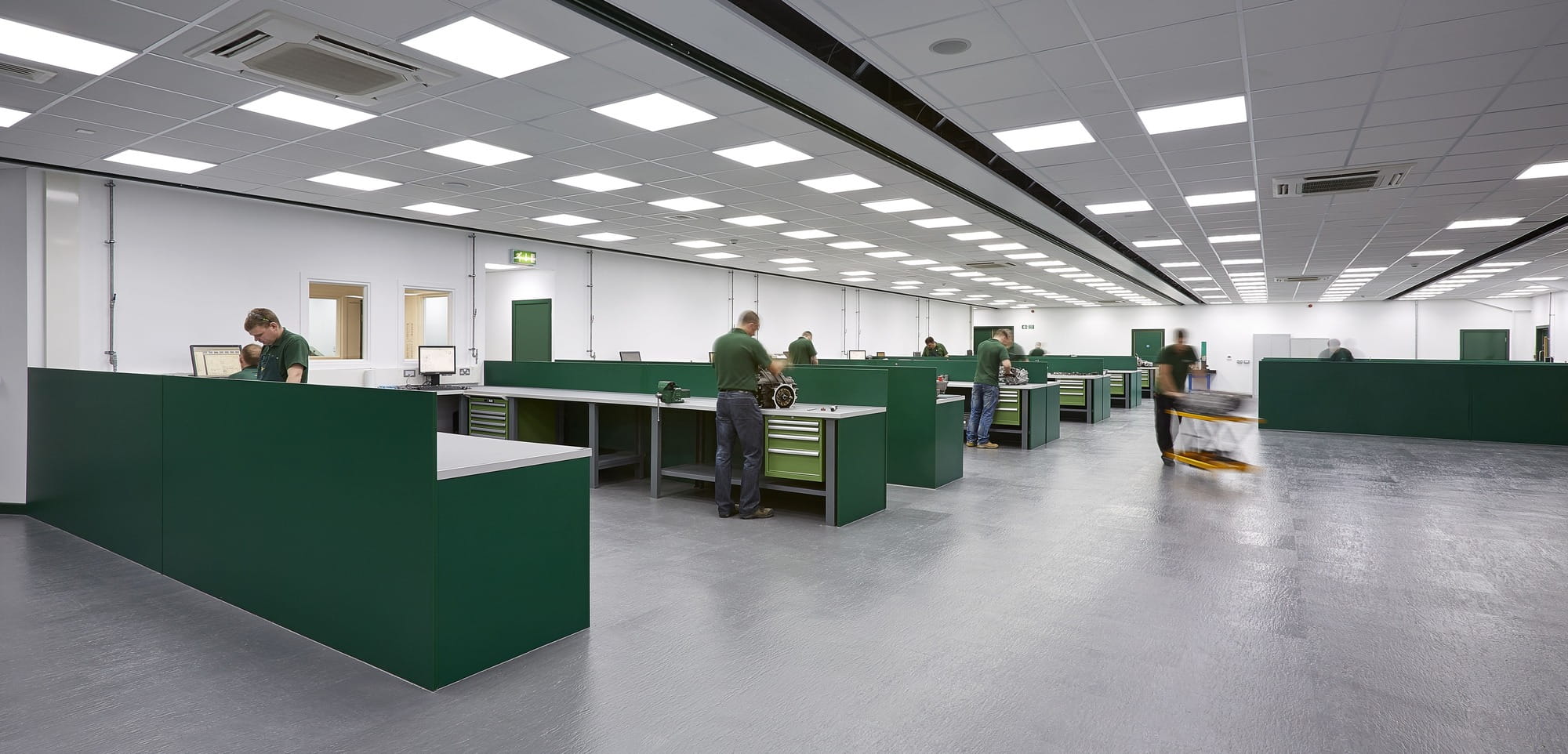 A spacious manufacturing workspace featuring several green workstations, with employees engaged in various tasks, under bright overhead lighting.