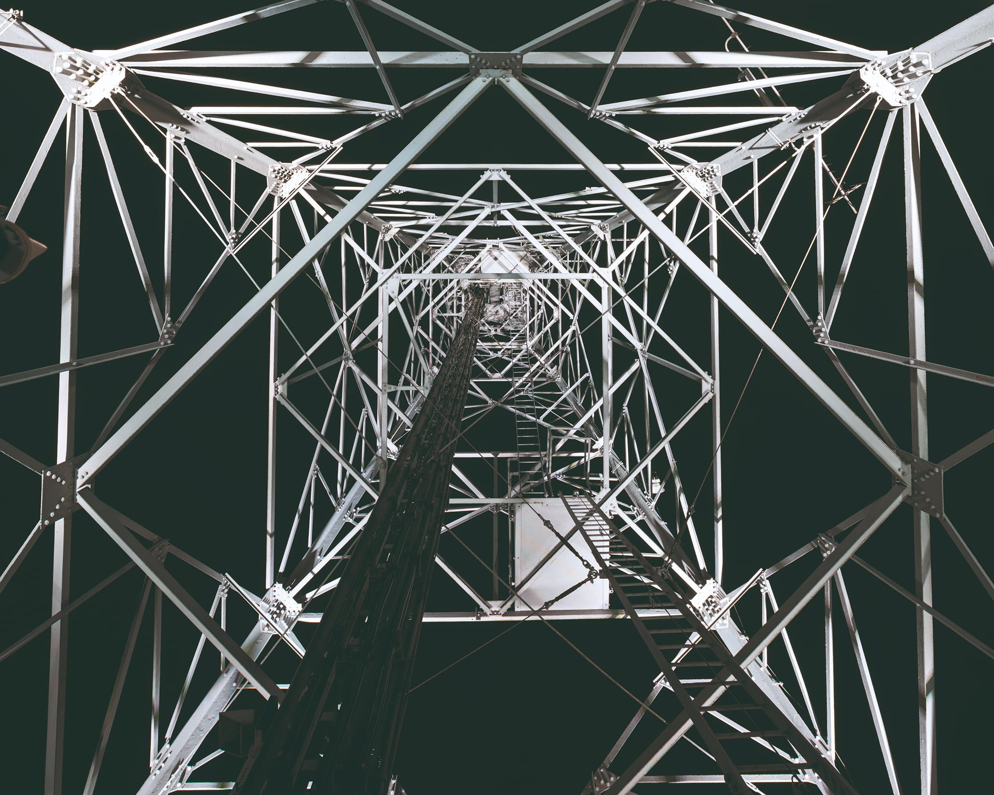 View looking up the interior of a tall metal structure, featuring a lattice framework and a vertical ladder against a dark background.