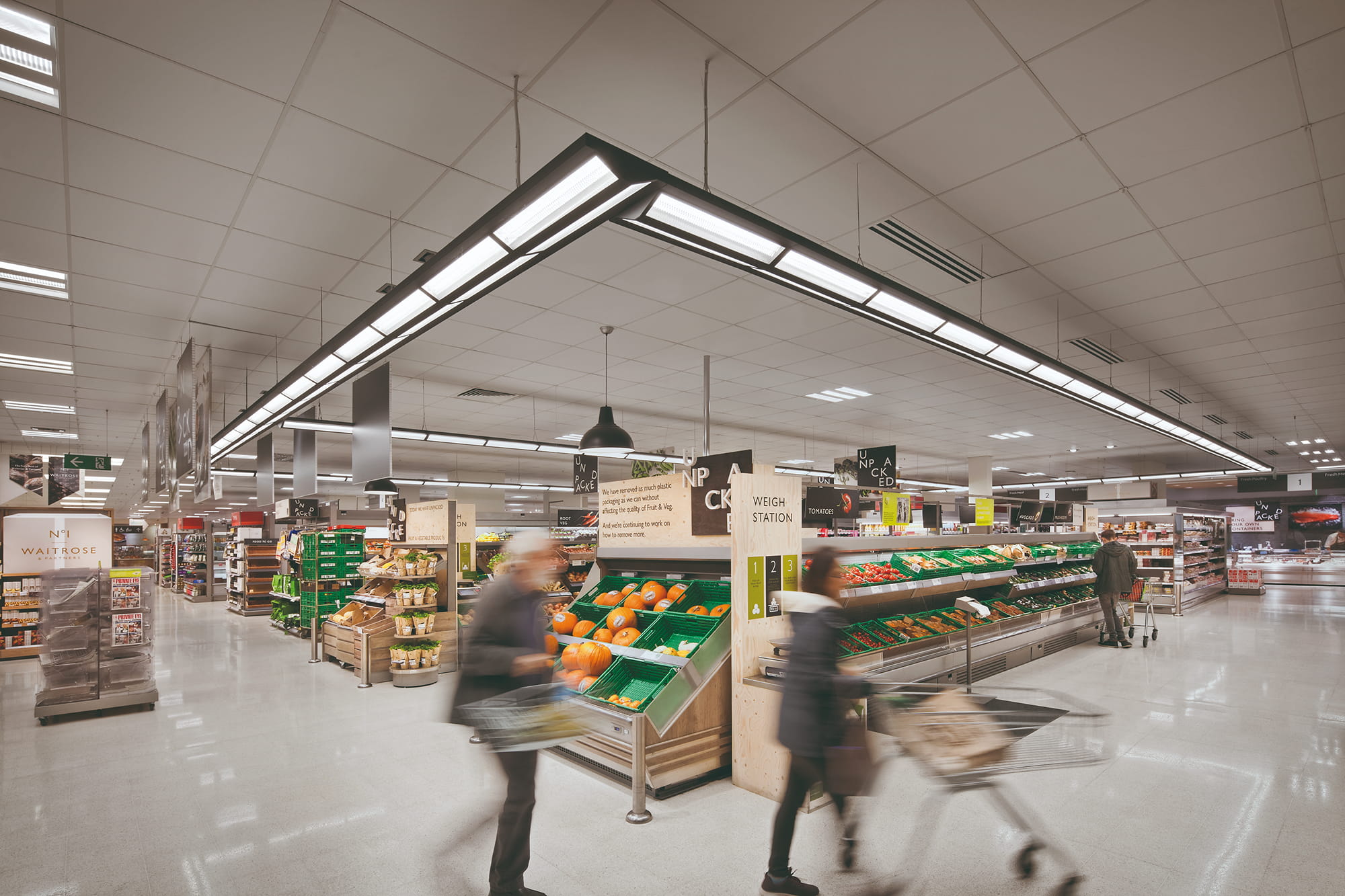Interior of a grocery store featuring produce displays, shopping carts, and customers browsing aisles under bright overhead lighting.