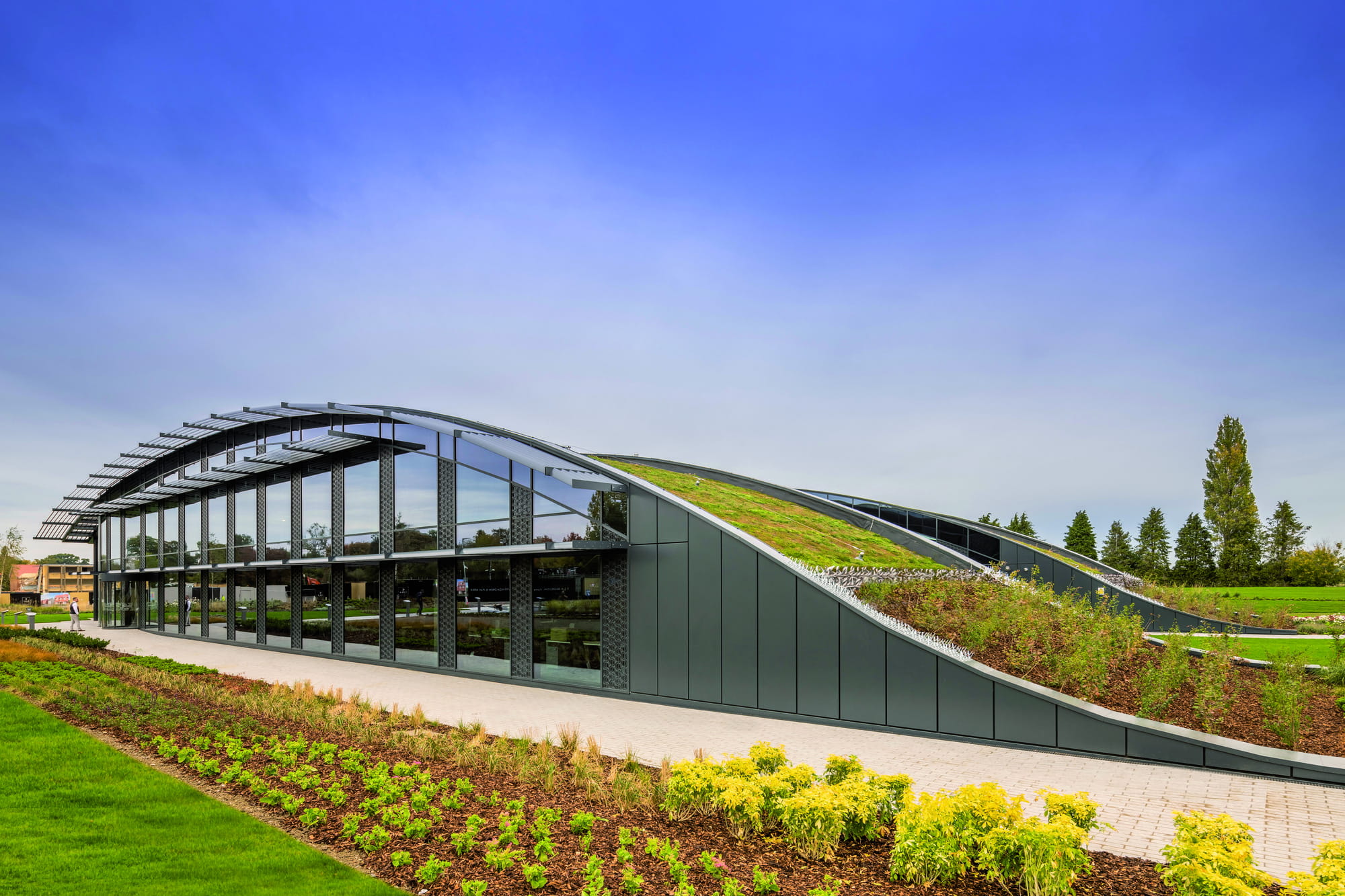 Modern building with a green roof and large glass windows, surrounded by landscaped gardens and a clear blue sky.