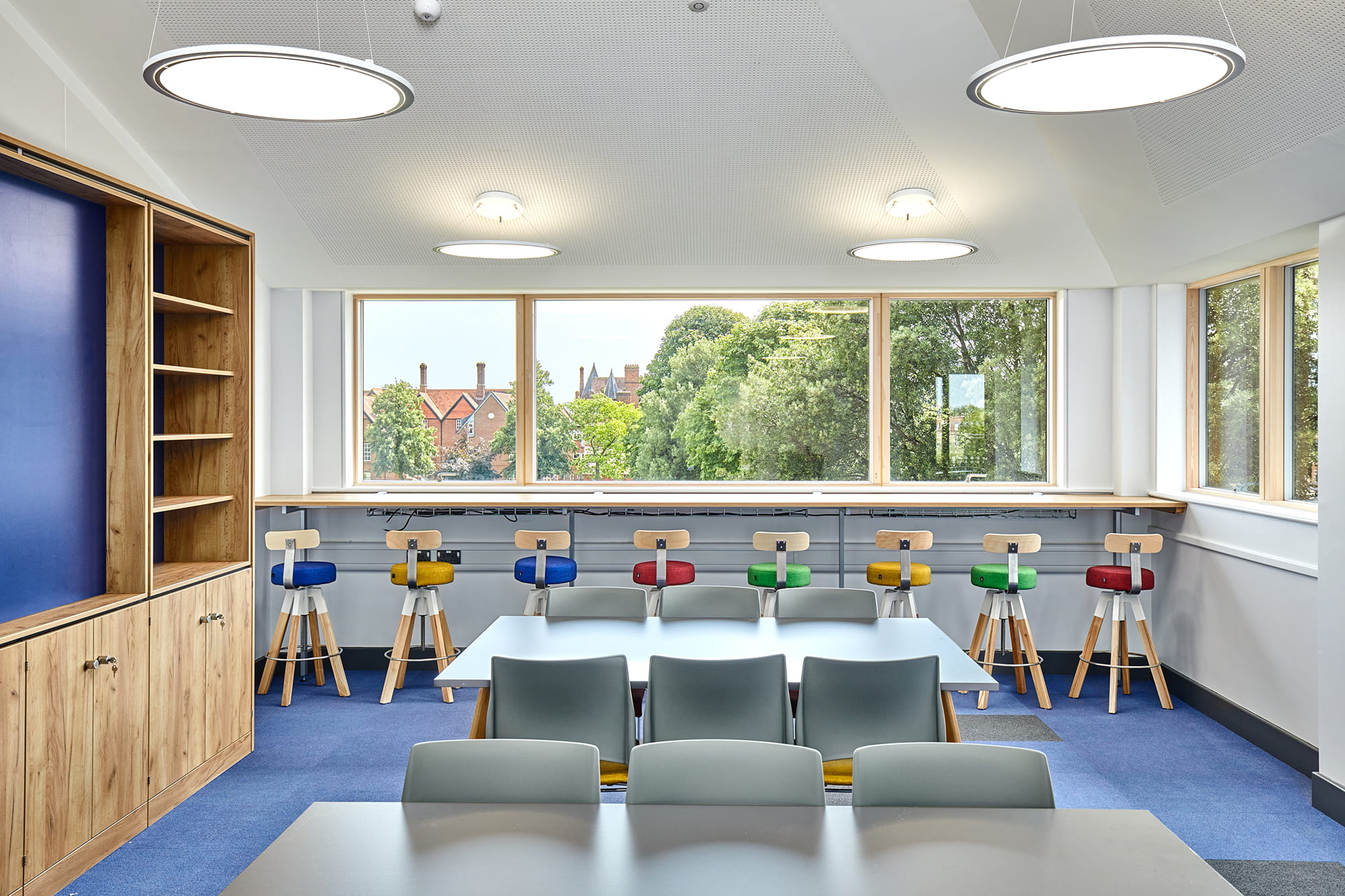 Bright classroom with large windows, featuring a long table, colorful stools, and wooden shelving.
