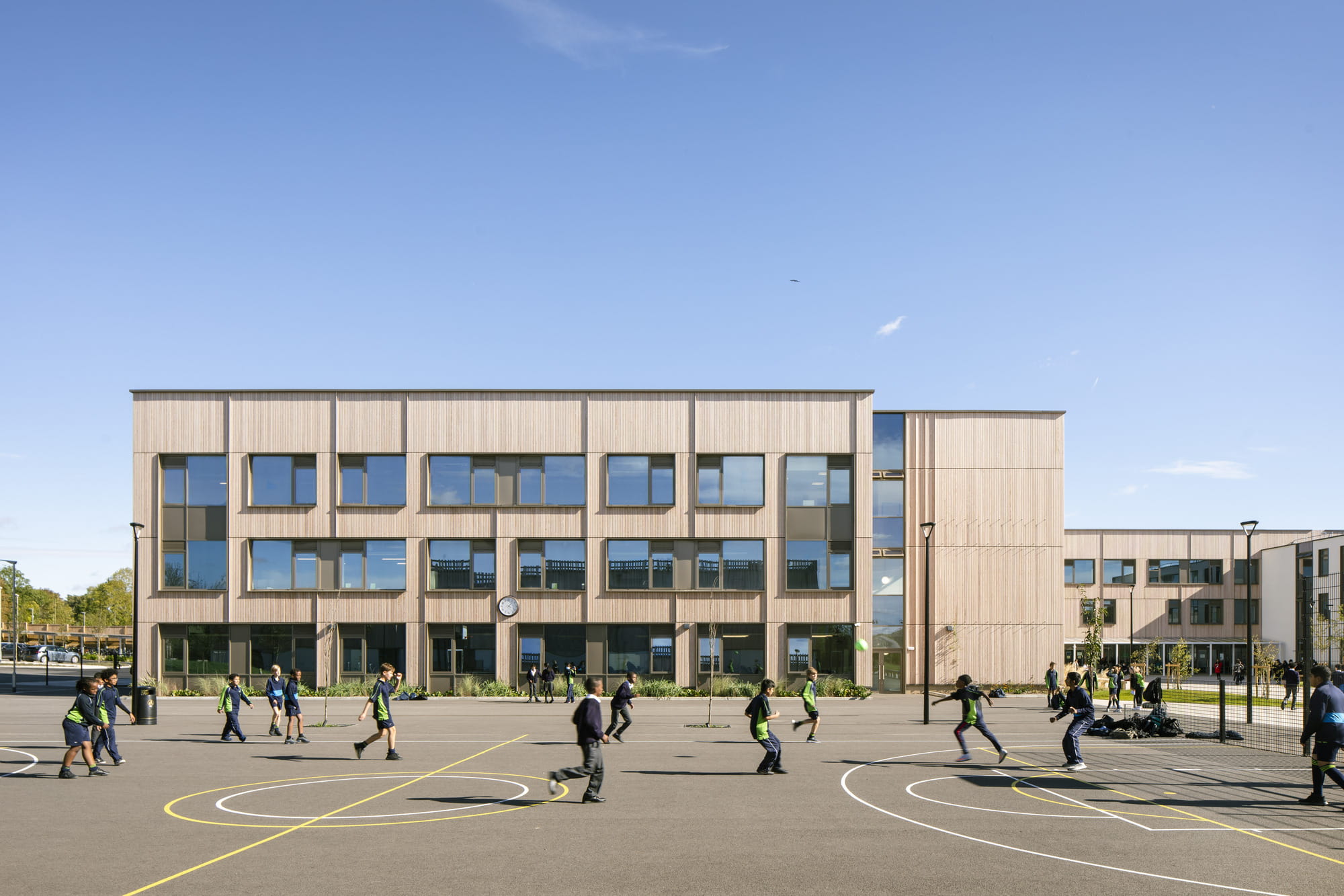 Modern school building with large windows and wooden exterior, surrounded by students playing on a basketball court.
