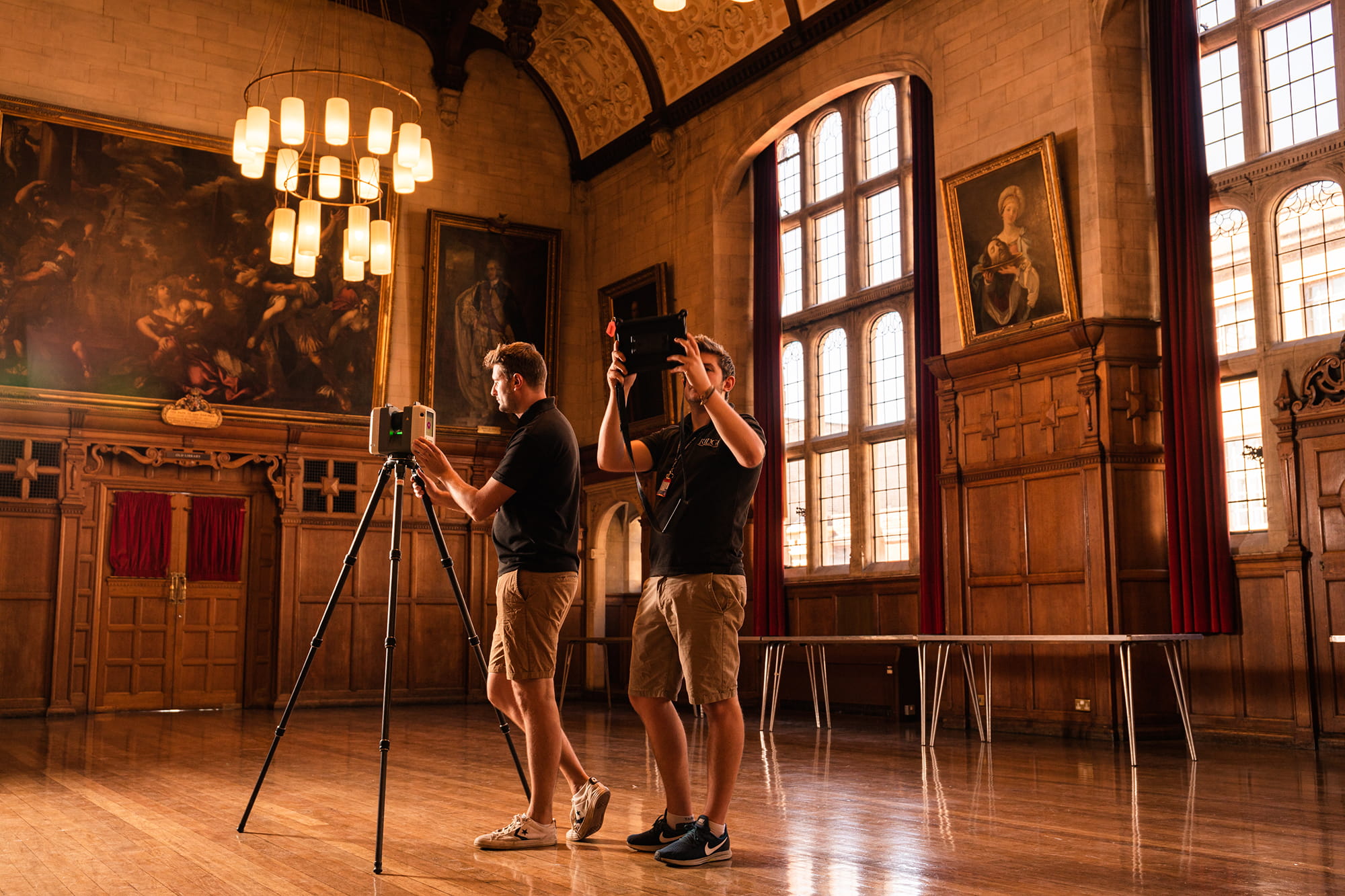 Two men using surveying equipment in a large, well-lit hall with wooden paneling and paintings on the walls.