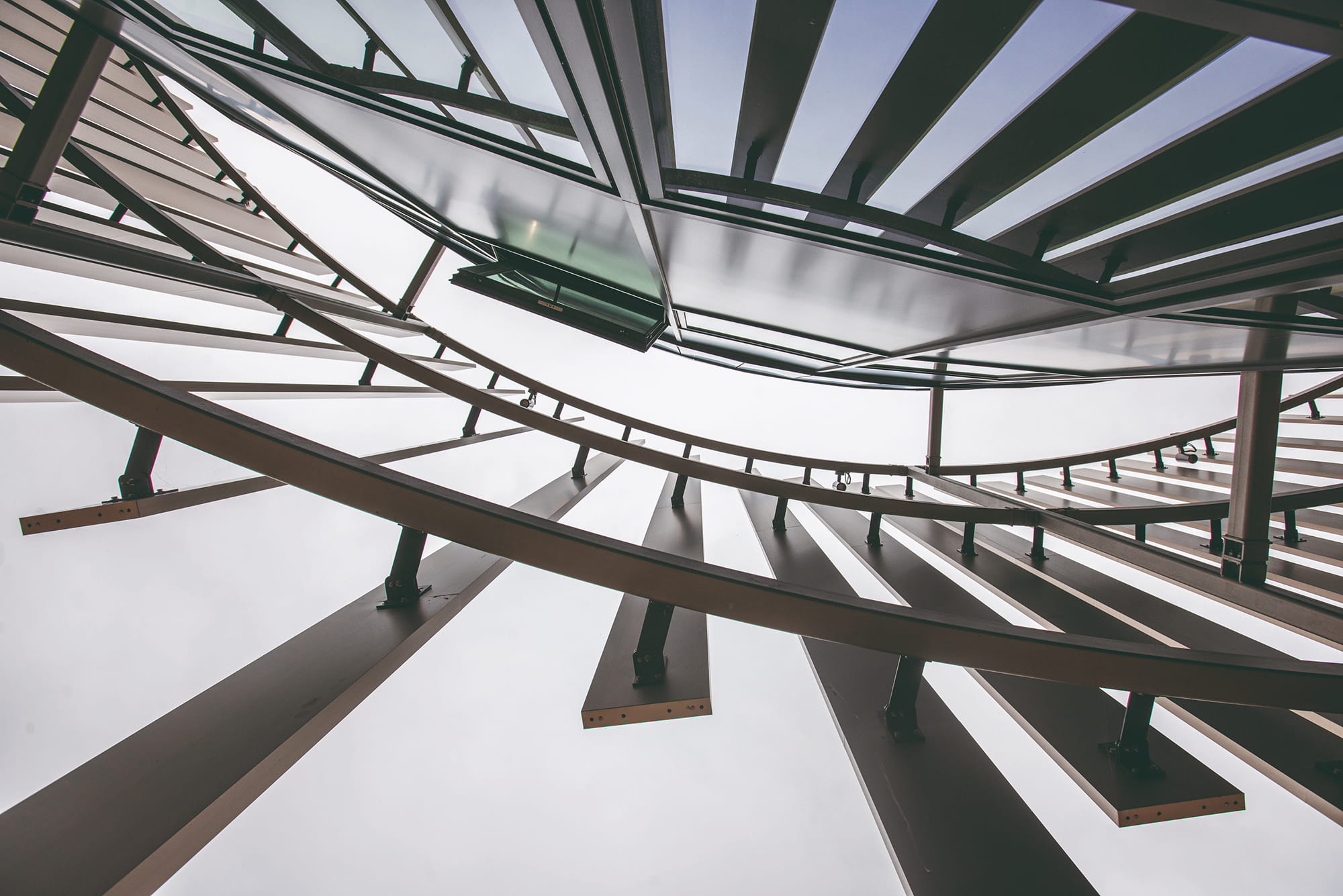 View of a modern architectural structure from below, featuring curved metal beams and glass panels against a cloudy sky.