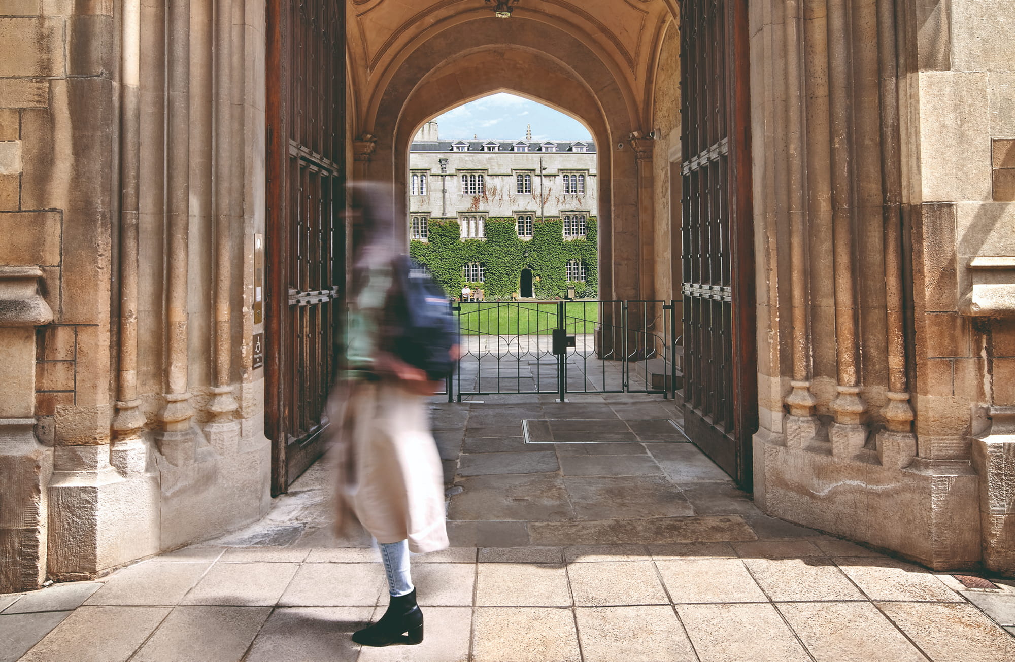 A blurred figure walks through an arched stone gateway, leading to a green courtyard surrounded by ivy-covered buildings.