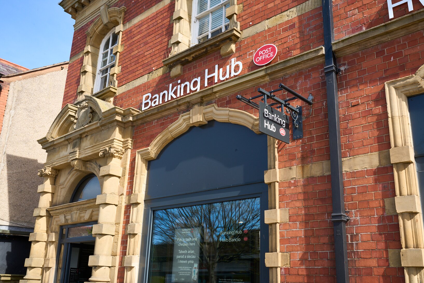 Exterior view of a Banking Hub building featuring a prominent sign and a Post Office logo, with decorative brickwork and a large window.