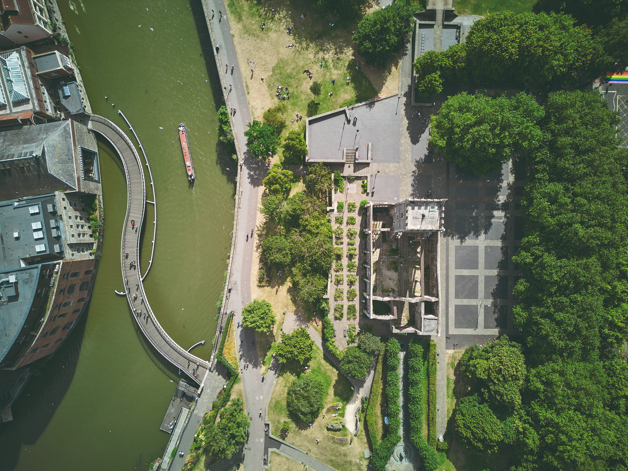 Aerial view of a green park area alongside a river, featuring a curved pedestrian bridge, a boat on the water, and remnants of a historic structure surrounded b