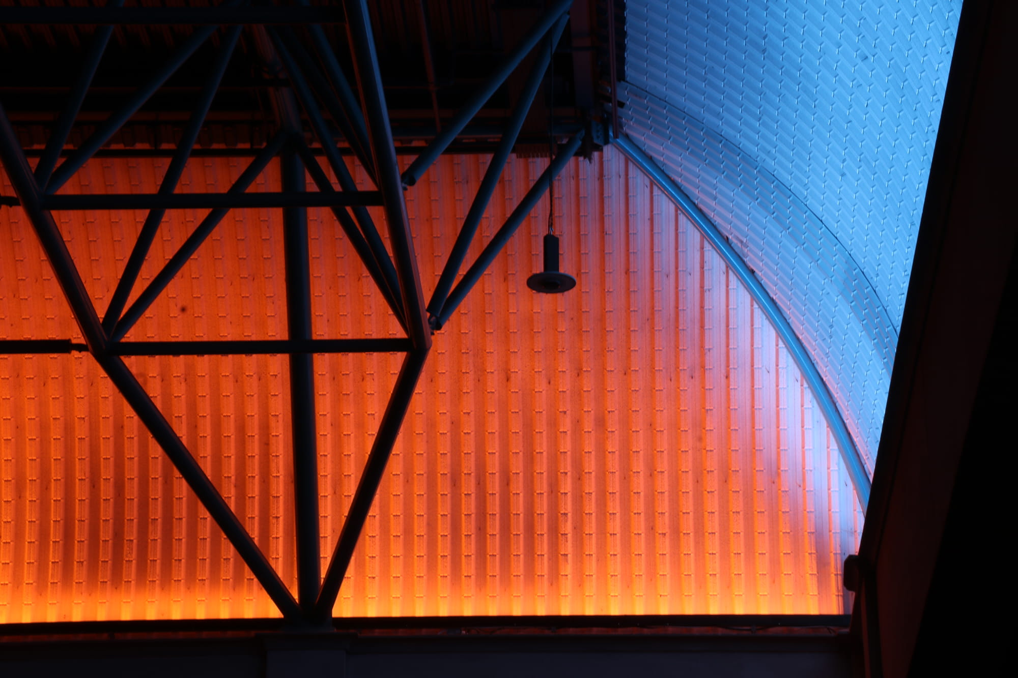 A ceiling with a triangular metal framework, featuring a gradient of orange and blue lighting on the textured wall panels.