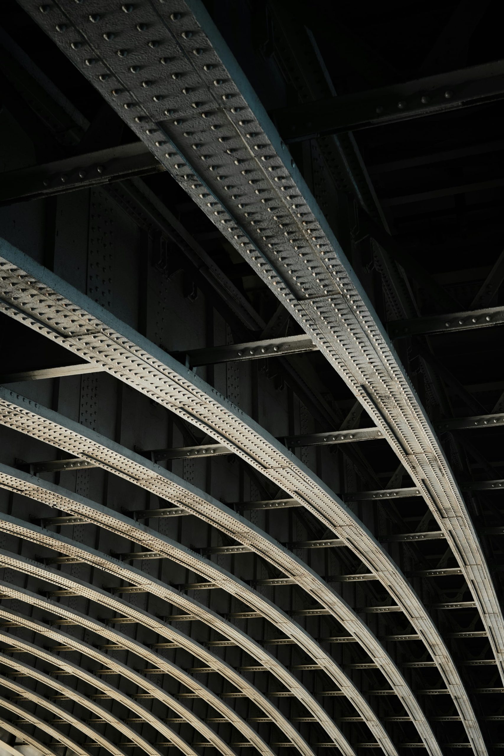 Curved steel beams and supports of a bridge, viewed from below, with a dark background.