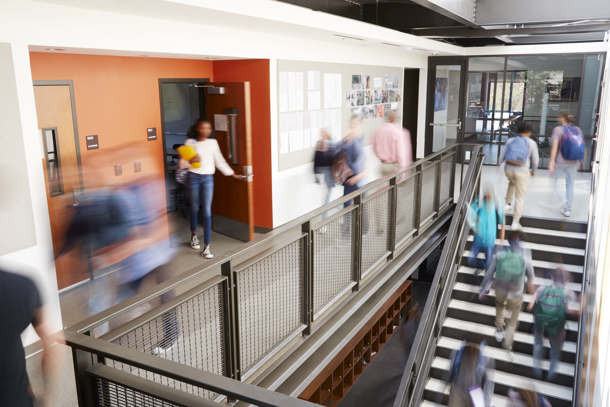 Students walking through a modern school hallway with open doors and a staircase.