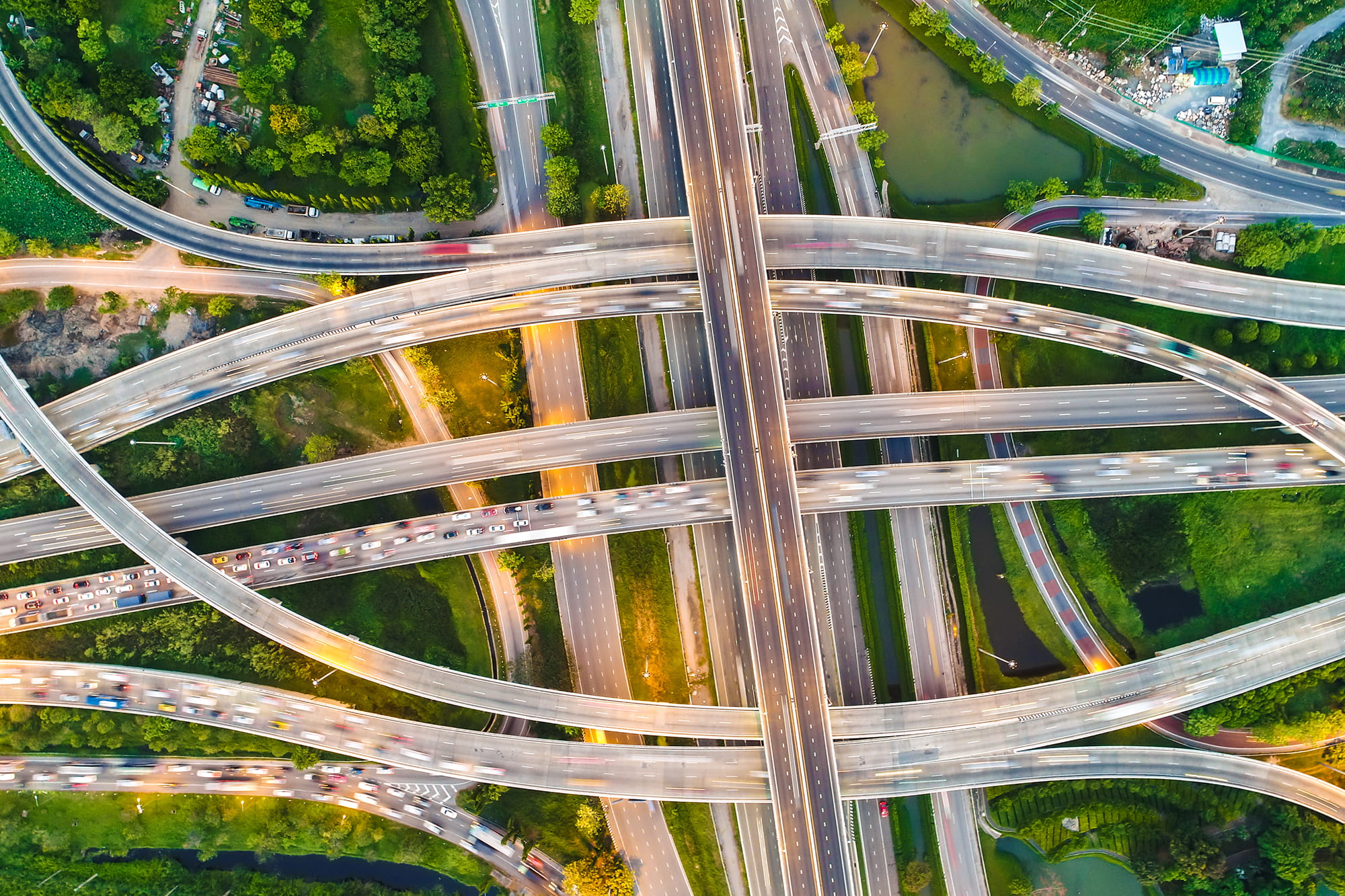 Aerial view of a complex highway interchange with multiple lanes and vehicles in motion, surrounded by greenery and roads.