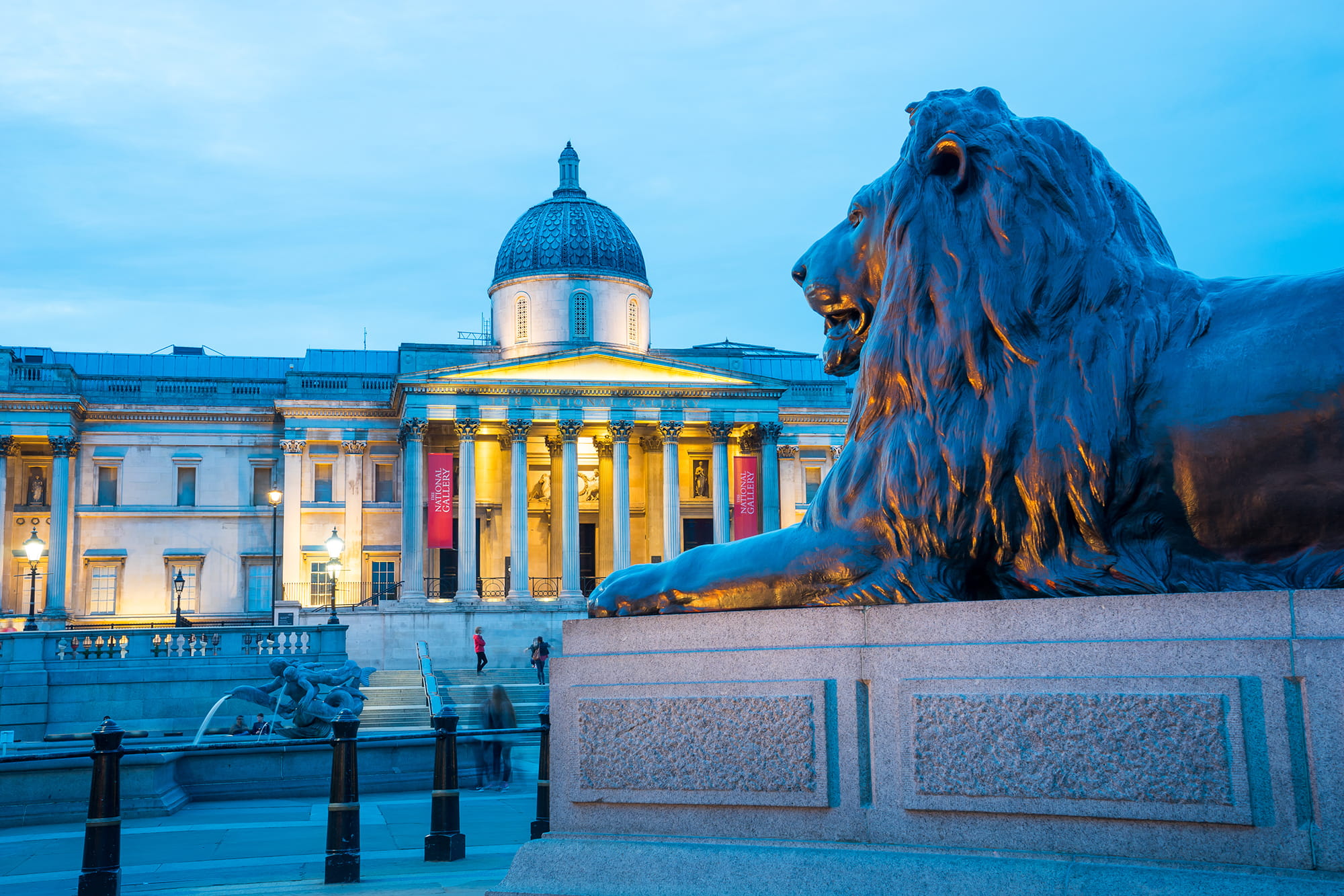 A bronze lion statue in the foreground with the National Gallery building illuminated in the background during twilight.