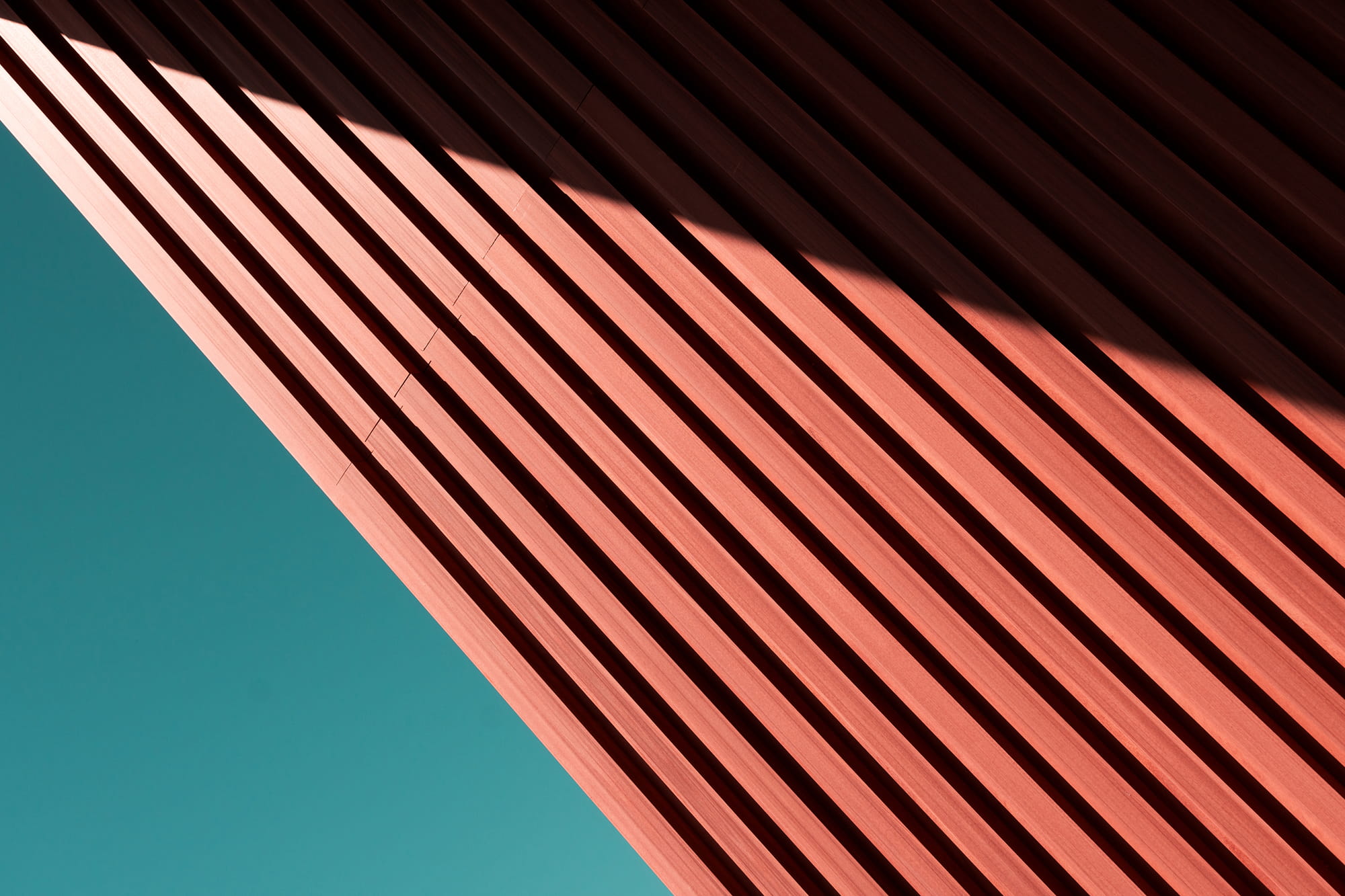 Diagonal wooden slats in a reddish hue against a turquoise background, creating a play of light and shadow.