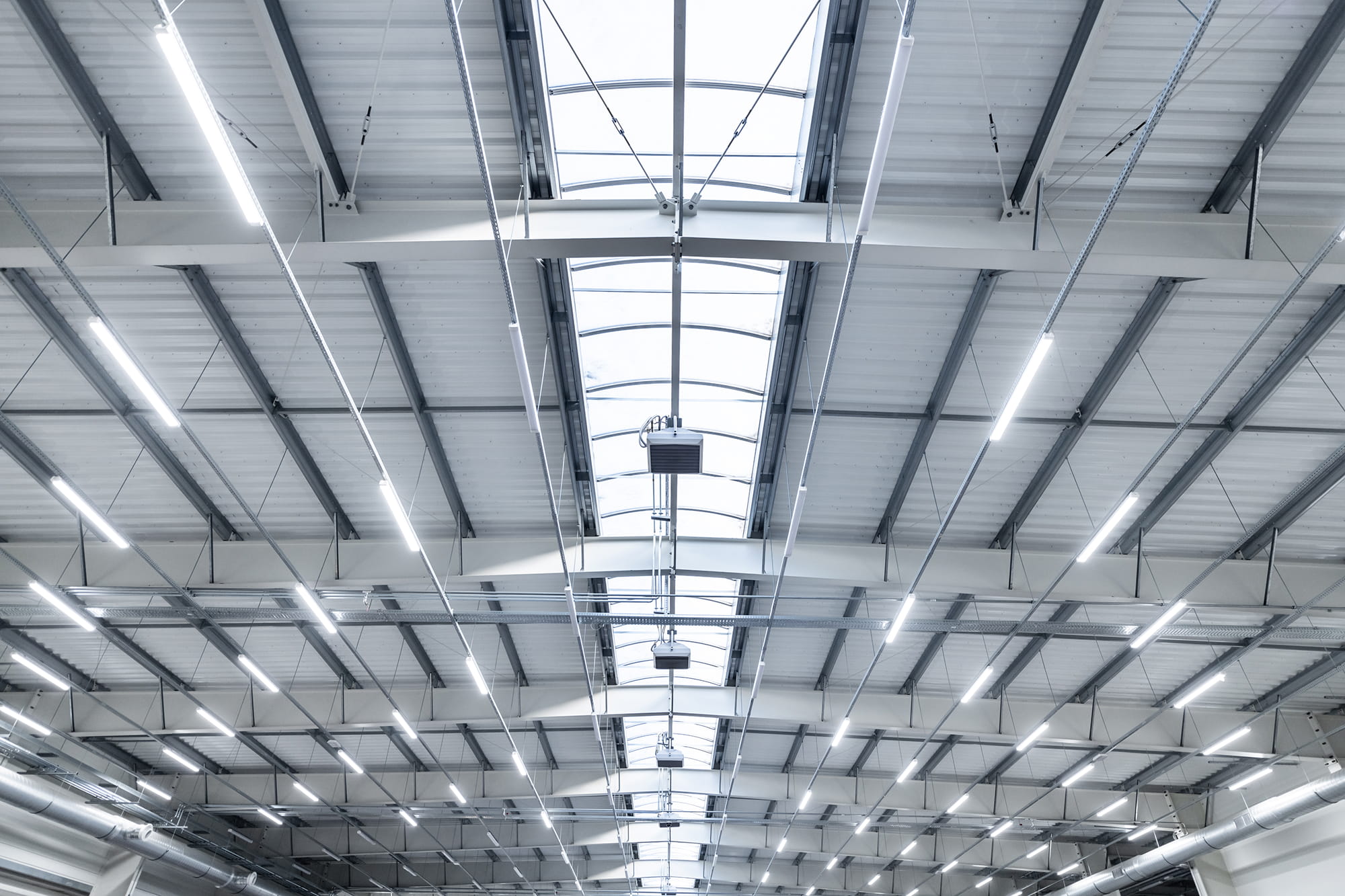 Ceiling of an industrial building featuring metal beams, skylights, and overhead lighting fixtures.