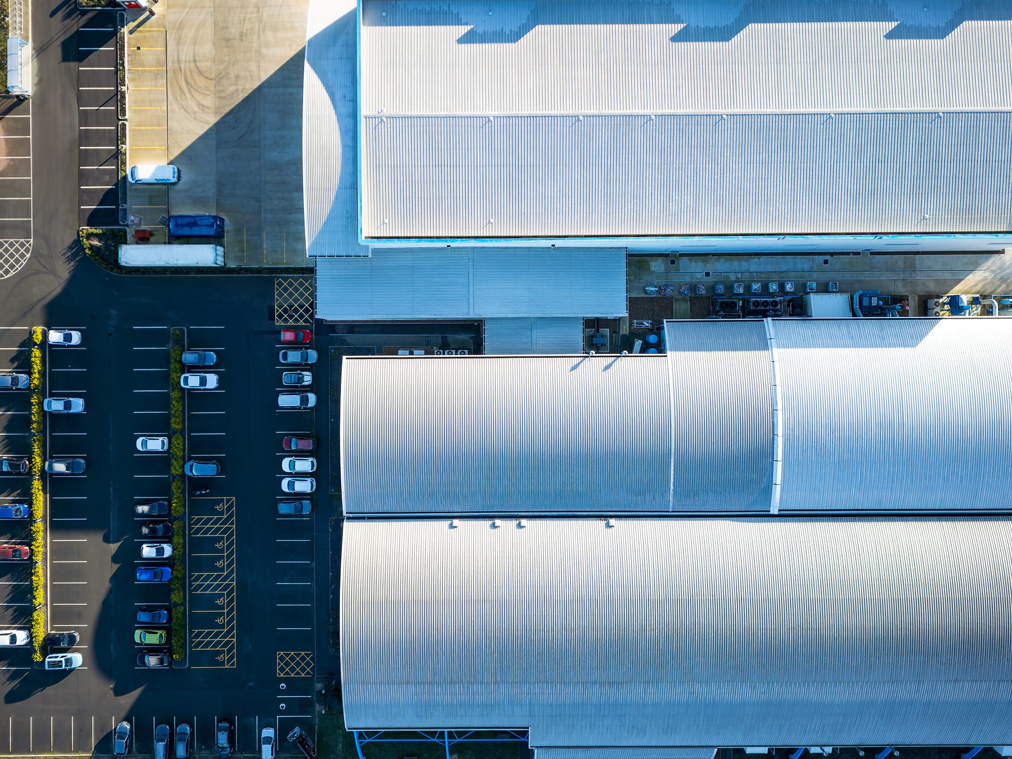 Aerial view of industrial buildings with corrugated roofs and a parking lot filled with vehicles.