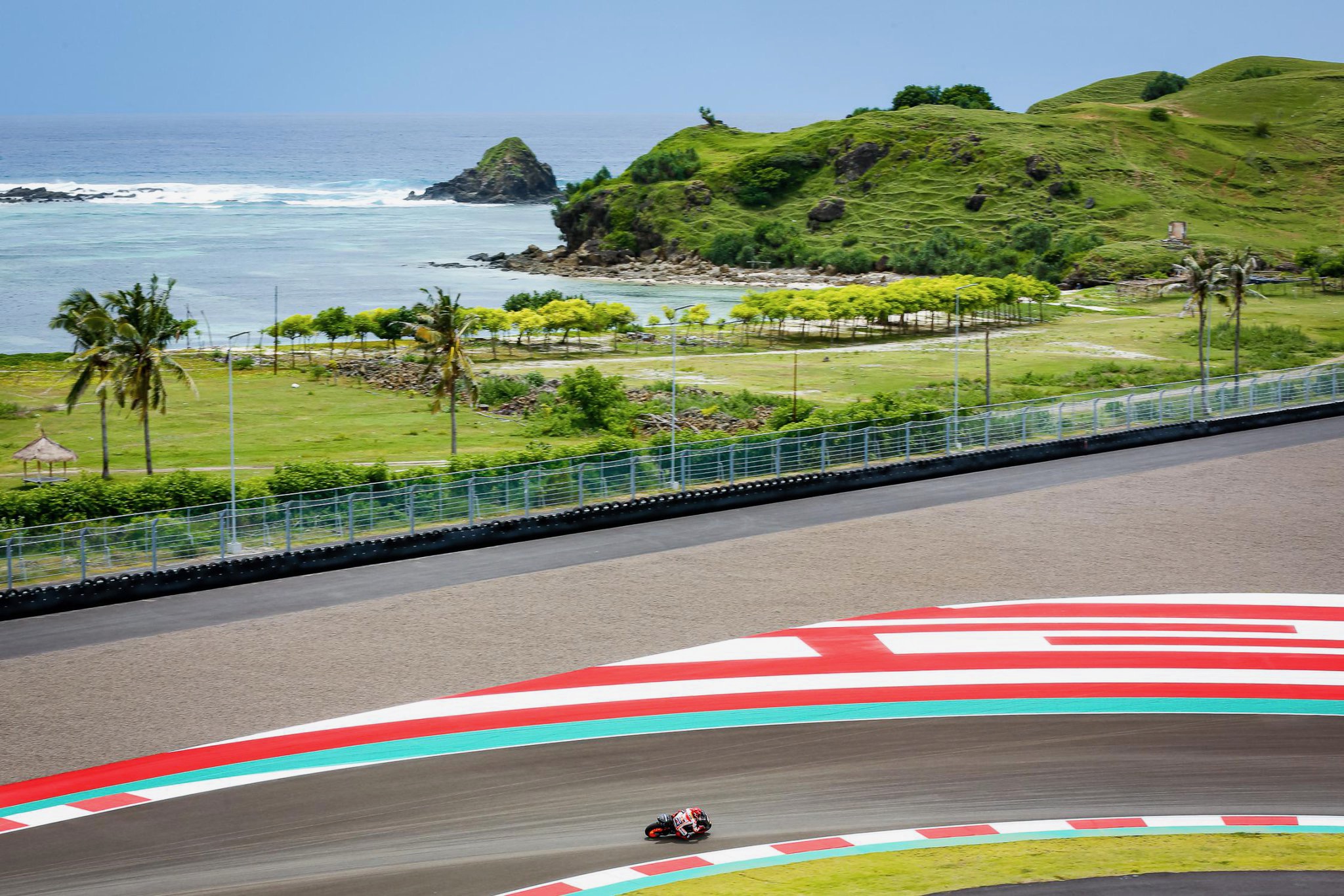 A motorcycle races around a curved track with a coastal landscape and hills in the background.