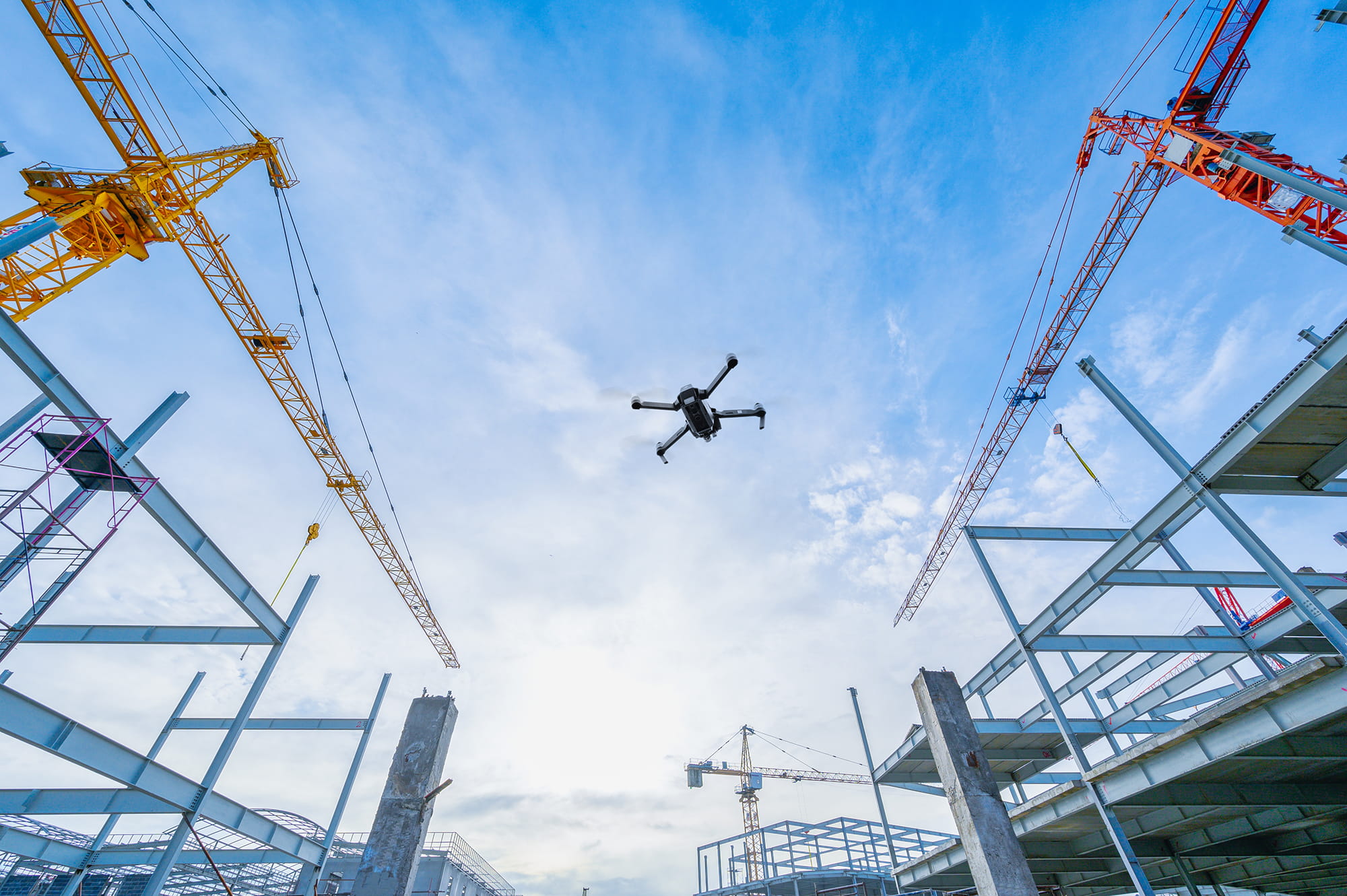 A drone hovers above a construction site with steel beams and cranes under a blue sky.