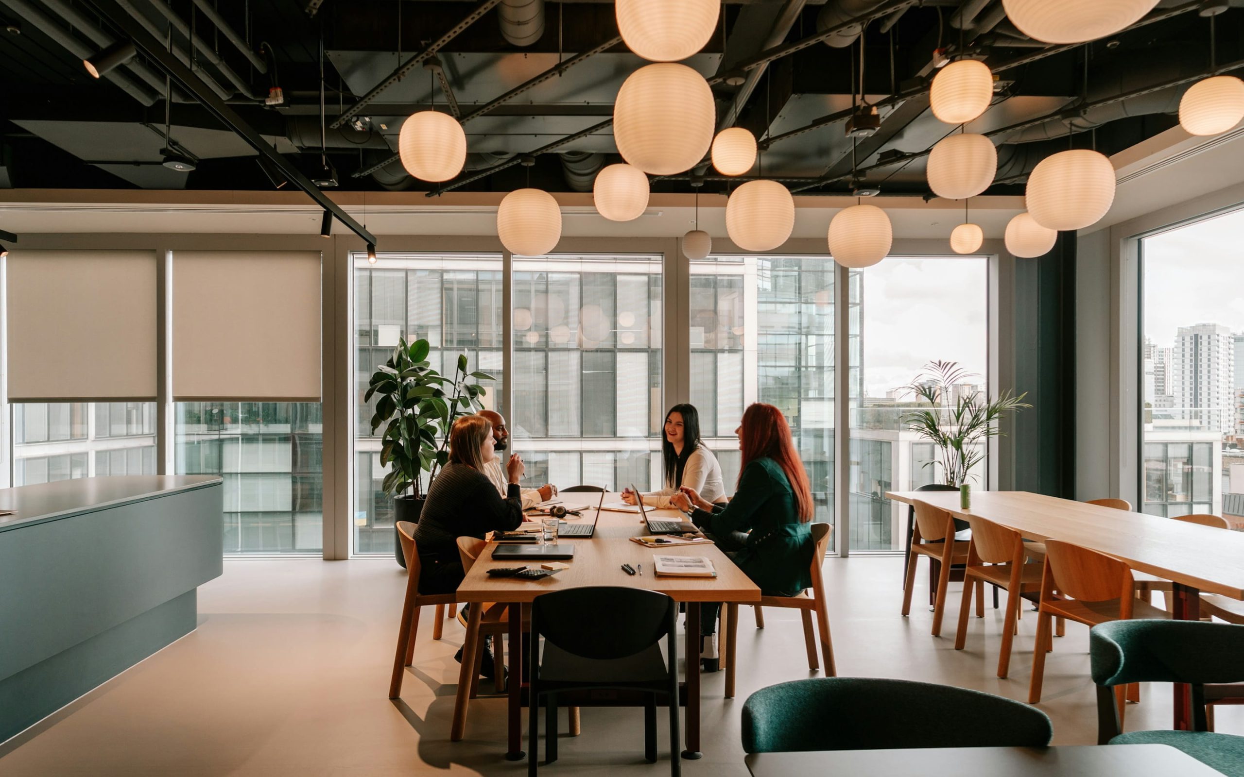 A group of four professionals engaged in a discussion around a wooden table in a modern office space with large windows and decorative pendant lights.