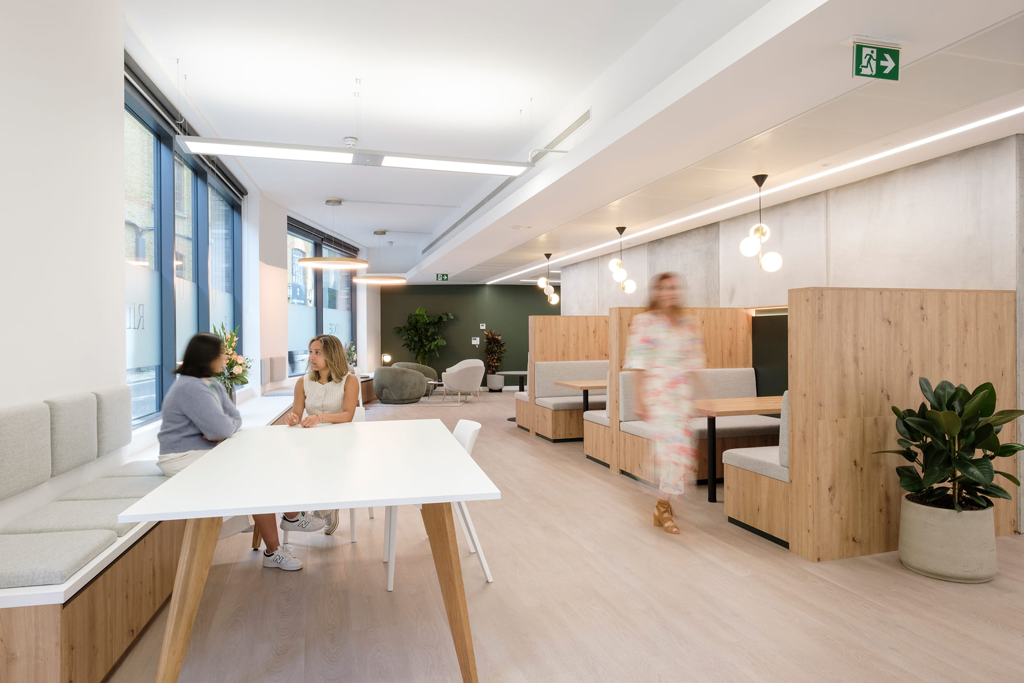 Modern office interior featuring a white table, wooden seating areas, and large windows, with two women engaged in conversation and a third person walking in th