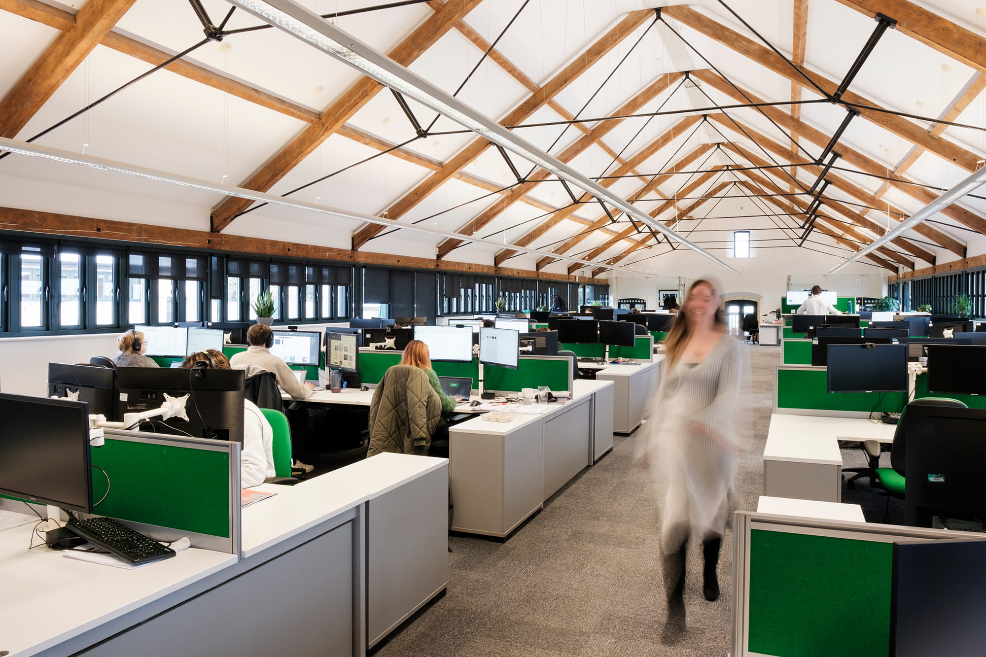 Open office space with multiple workstations, computers, and employees engaged in work, featuring a high wooden-beamed ceiling.