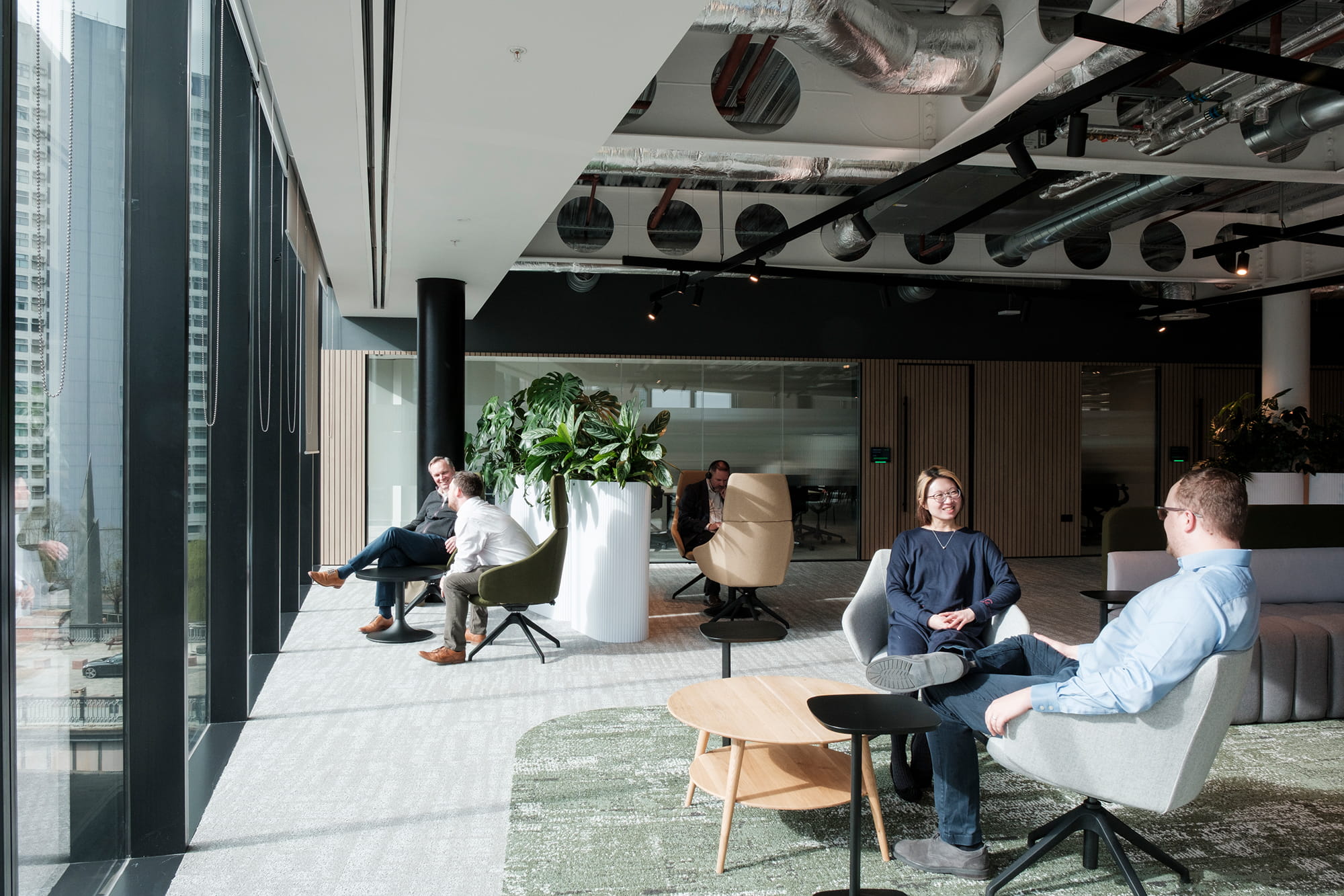 Modern office interior featuring several people engaged in conversation, large windows allowing natural light, and greenery in the background.