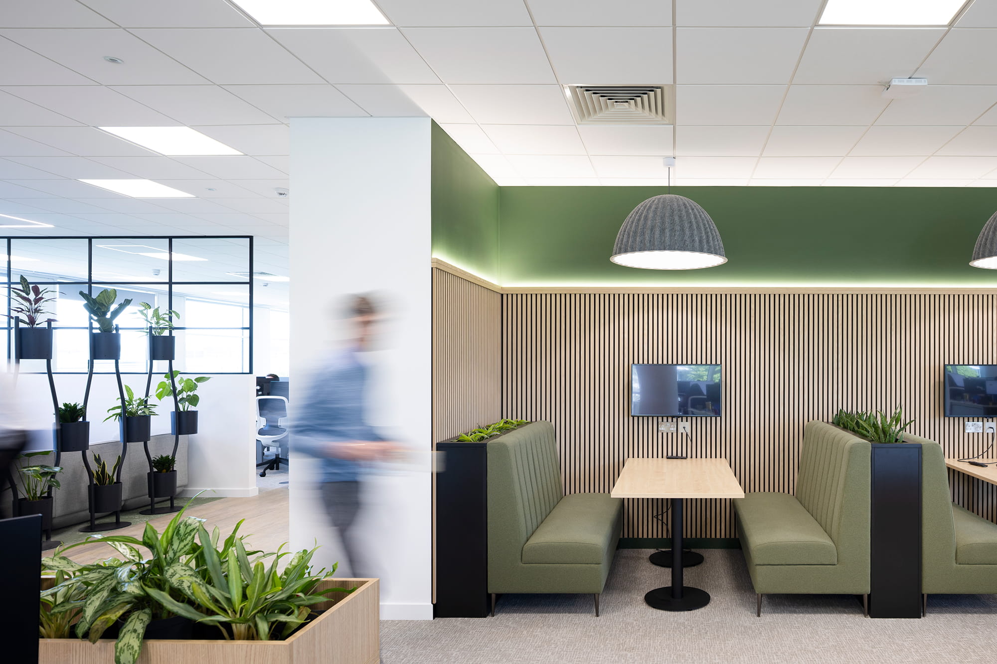 Modern office interior featuring green seating booths, wooden paneling, and potted plants, with a blurred figure walking past.