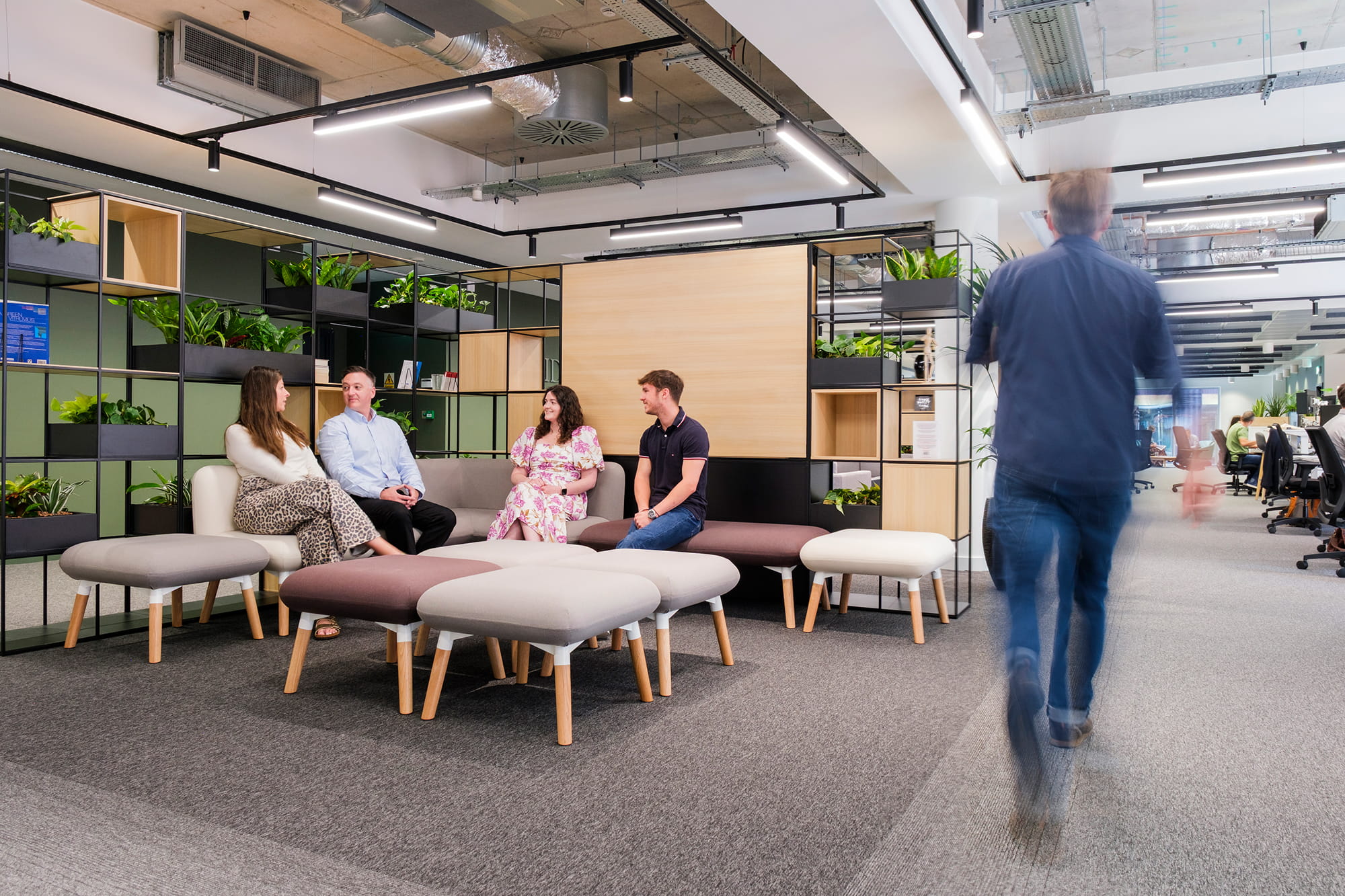 A group of four people engaged in conversation while seated on modern benches in a contemporary office space with greenery and open workstations in the backgrou