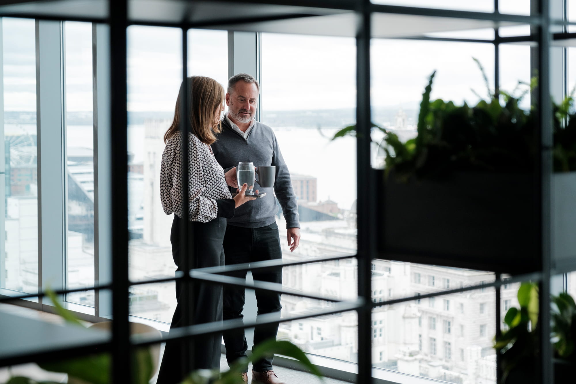 Two people stand by a large window in an office, holding drinks and looking out at the cityscape of Liverpool.
