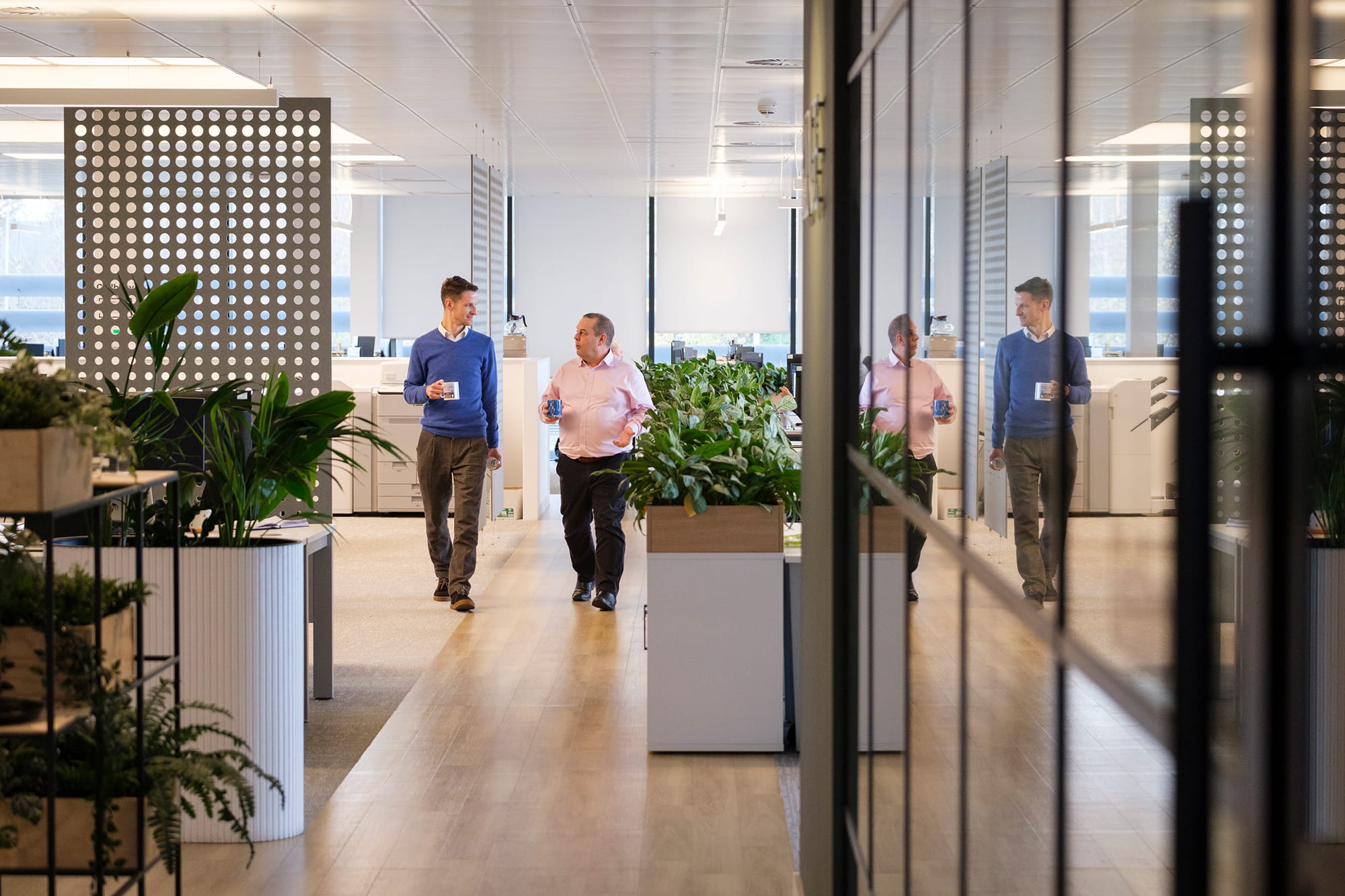 Two men walk through a modern office space with plants, one holding a coffee mug and the other a glass of water.