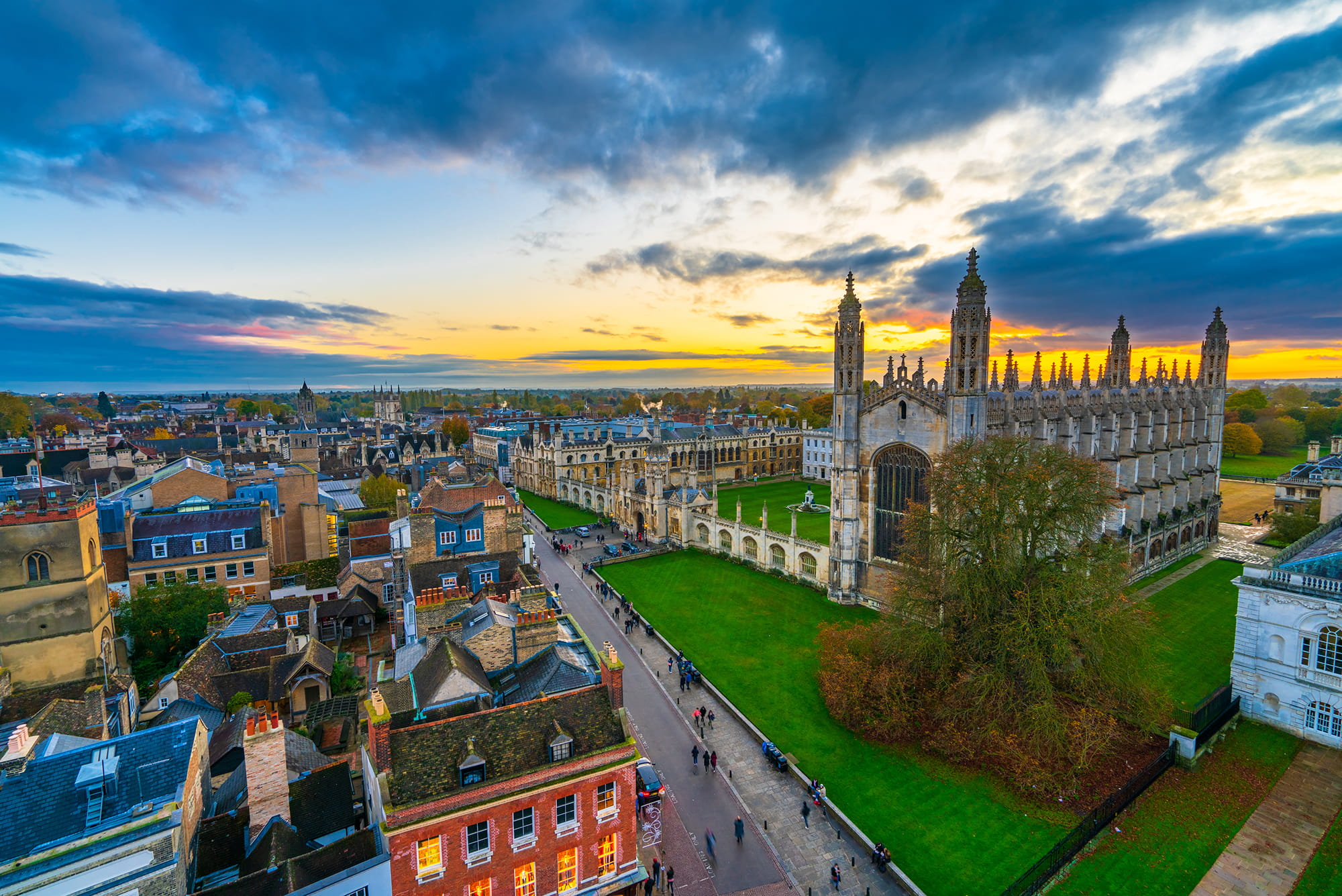 Aerial view of Cambridge at sunset, featuring historic buildings, rooftops, and green spaces.
