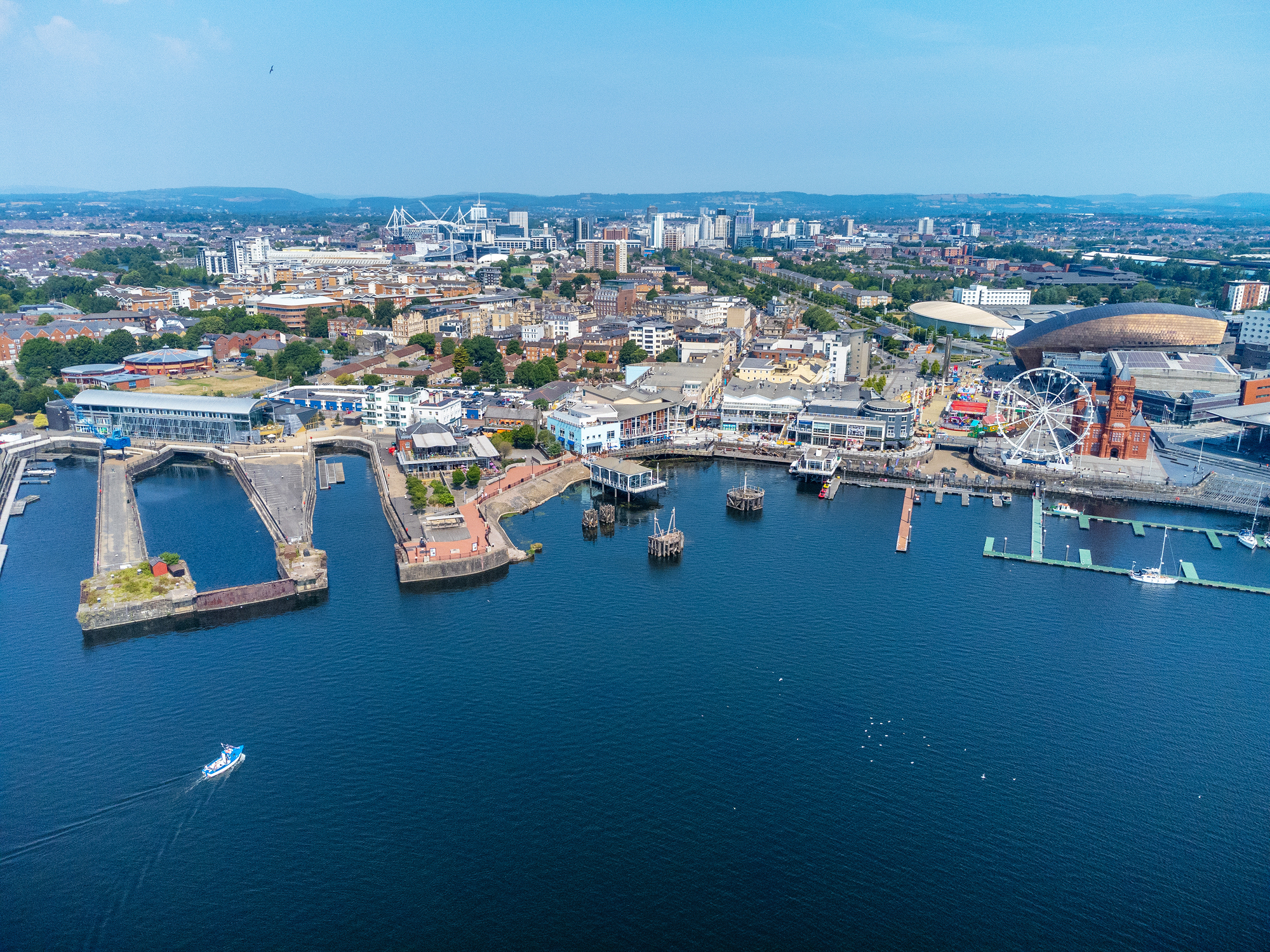 Aerial view of Cardiff's waterfront, featuring the bay, docks, and city skyline with various buildings and a Ferris wheel.
