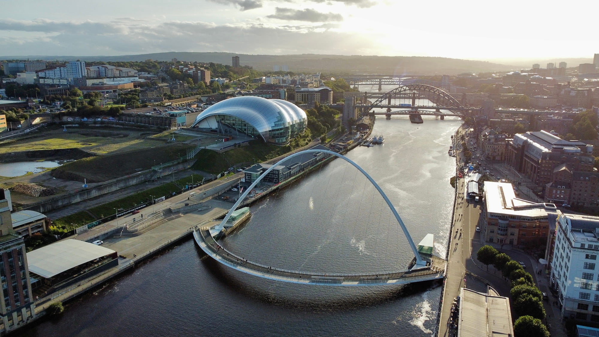 Aerial view of the Quayside in Newcastle, featuring the Sage Gateshead concert hall, the Millennium Bridge, and the River Tyne with city buildings in the backgr