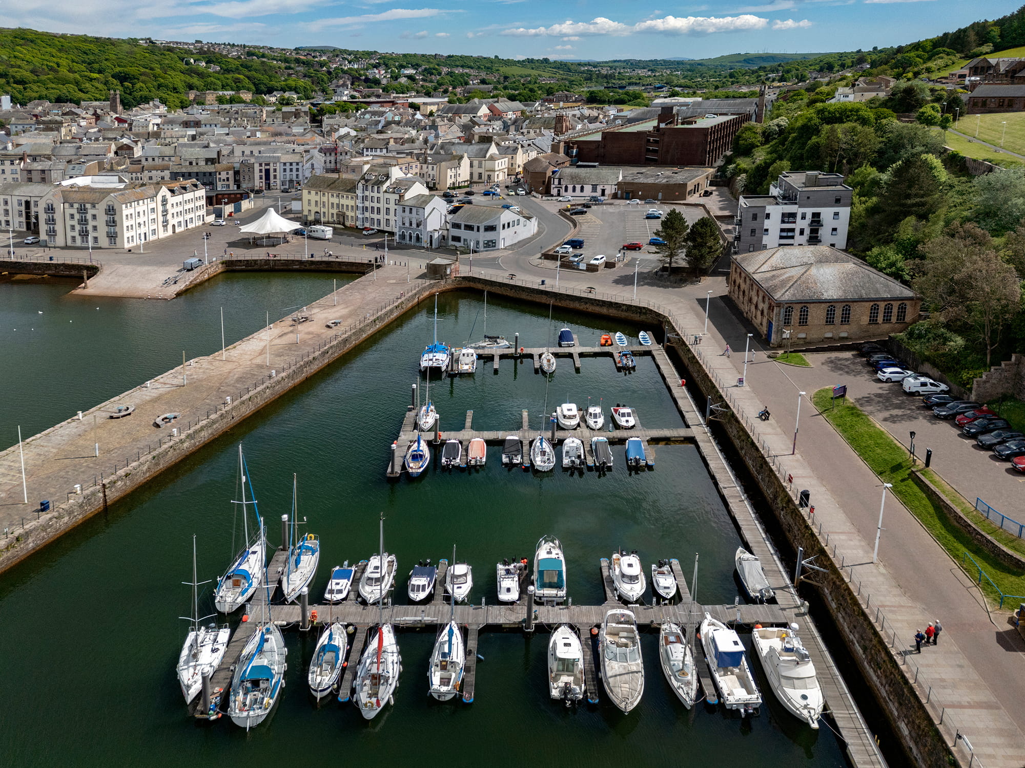 Aerial view of a marina in Whitehaven, featuring several boats docked at the pier, surrounded by buildings and green hills in the background.