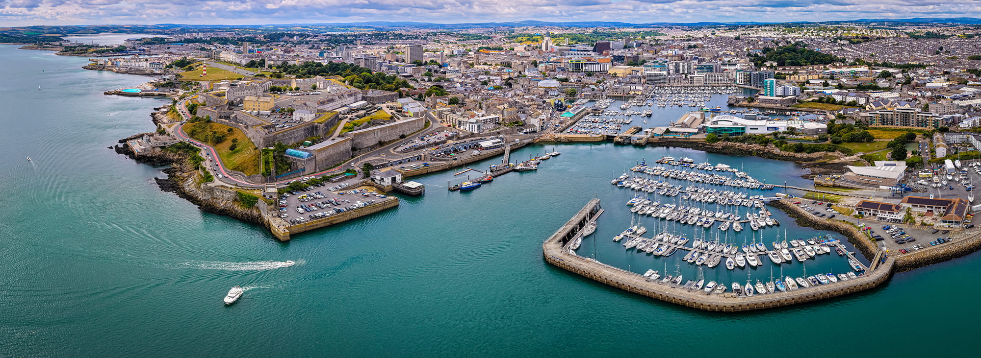 Aerial view of Plymouth harbor featuring boats, marinas, and cityscape with hills in the background.