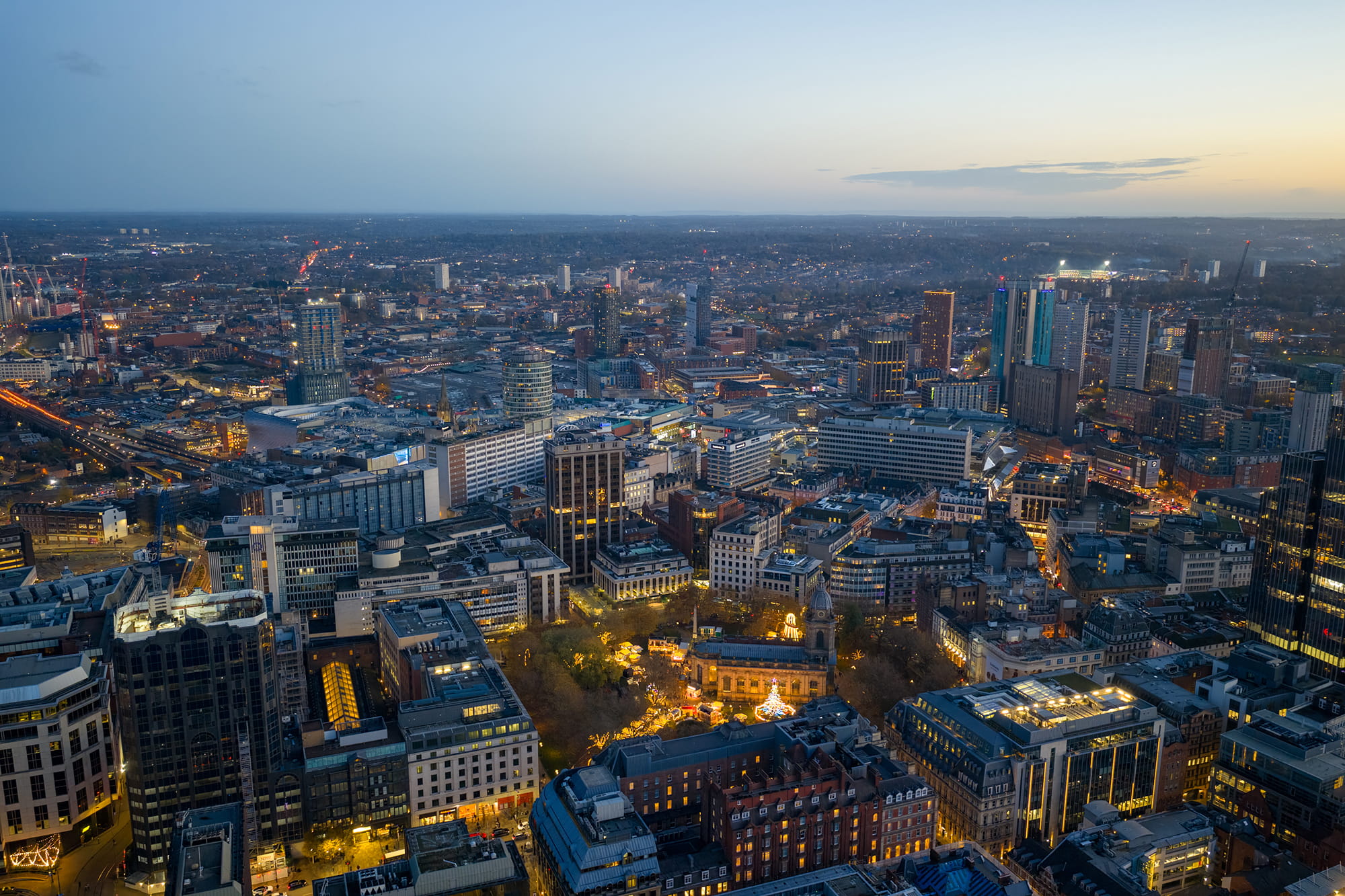 Aerial view of Birmingham city center at dusk, showcasing a mix of modern and historic buildings illuminated by city lights.