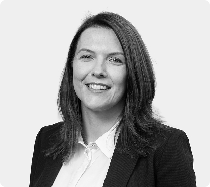Professional headshot of a woman with shoulder-length brown hair, wearing a black blazer and white shirt, smiling against a light gray background.