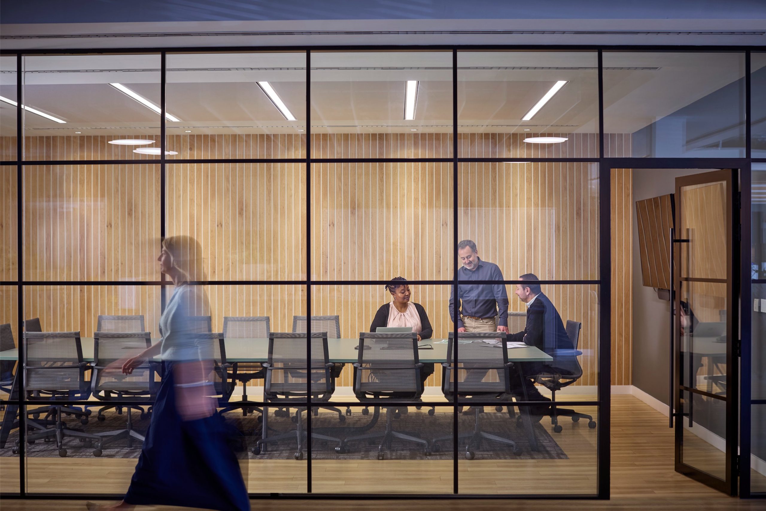 A woman walks past a glass-walled conference room where three people are engaged in discussion around a table.