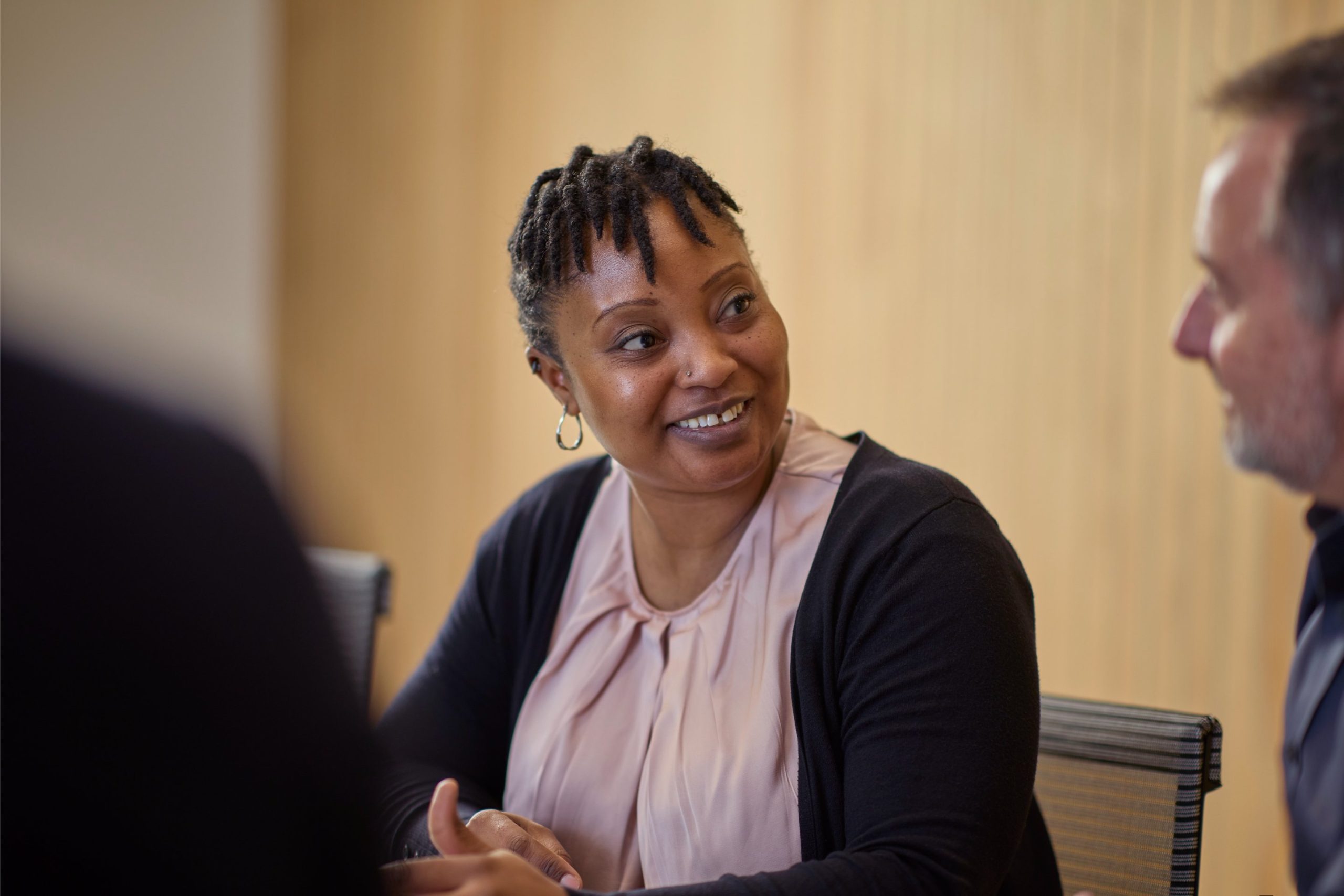 A woman with short, coiled hair and earrings smiles while engaging in conversation with a man in a meeting setting.