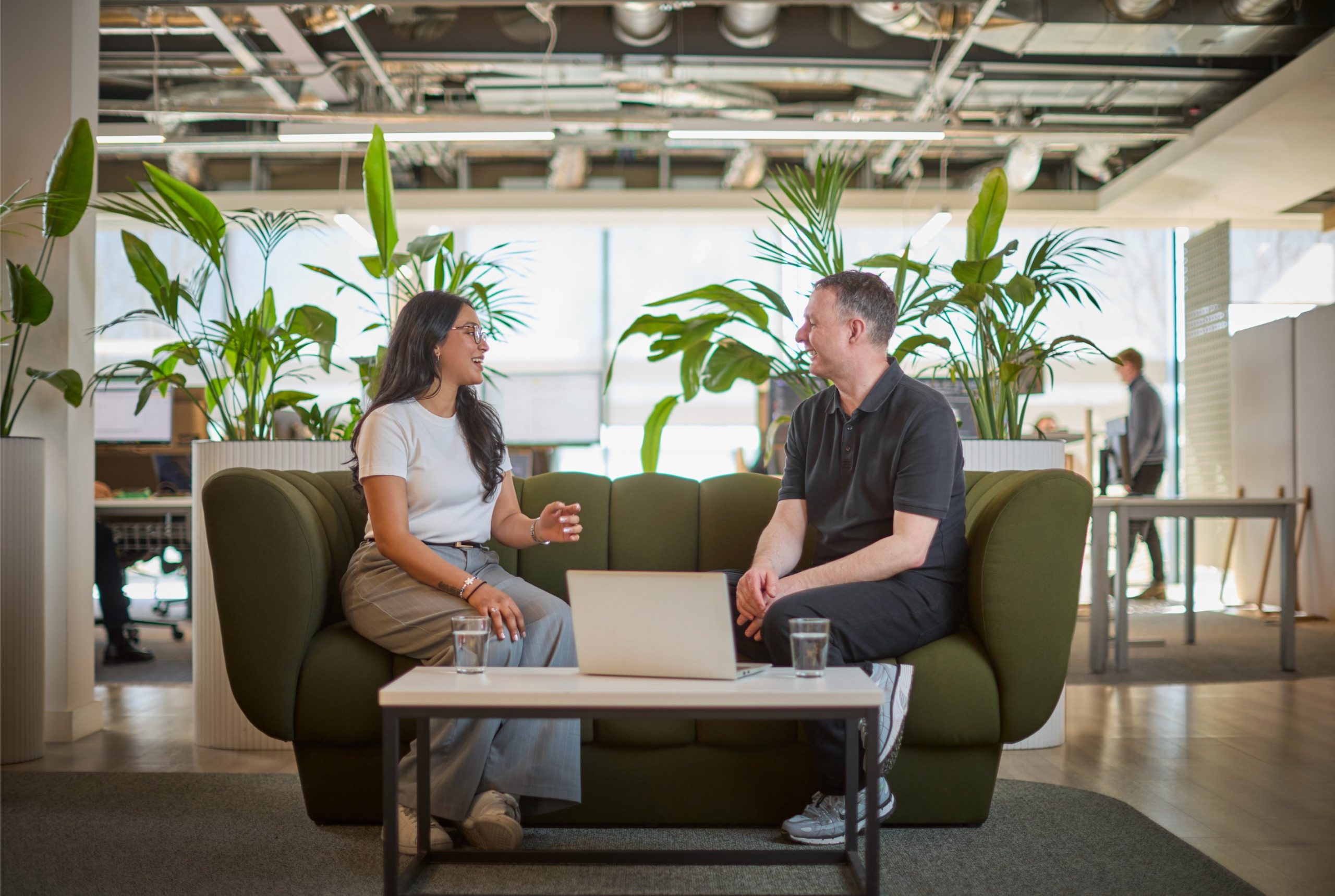 Two people engaged in conversation on a green sofa in a modern office space, surrounded by plants, with a laptop and glasses of water on a table.