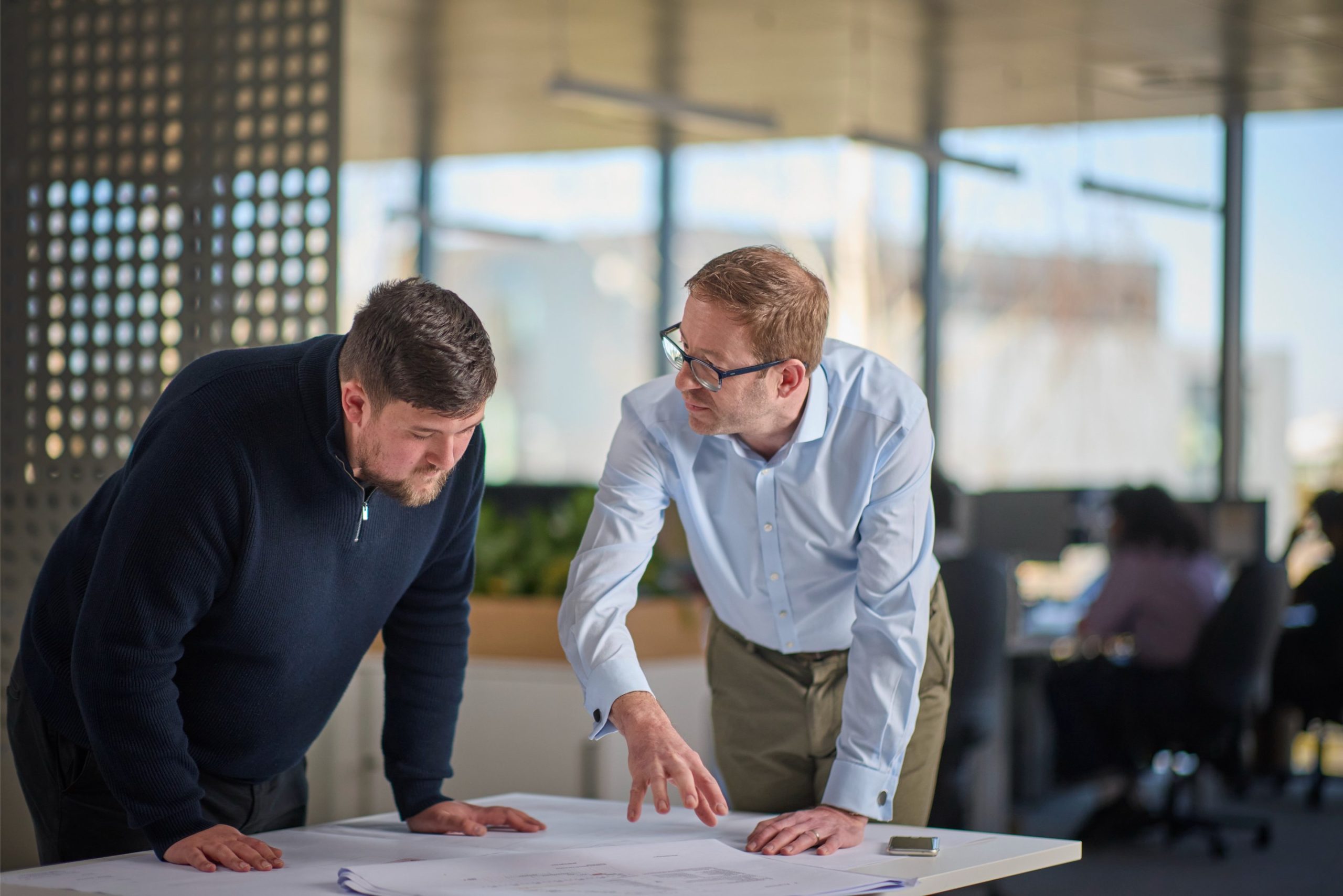 Two men discuss architectural plans while leaning over a table in a modern office setting.