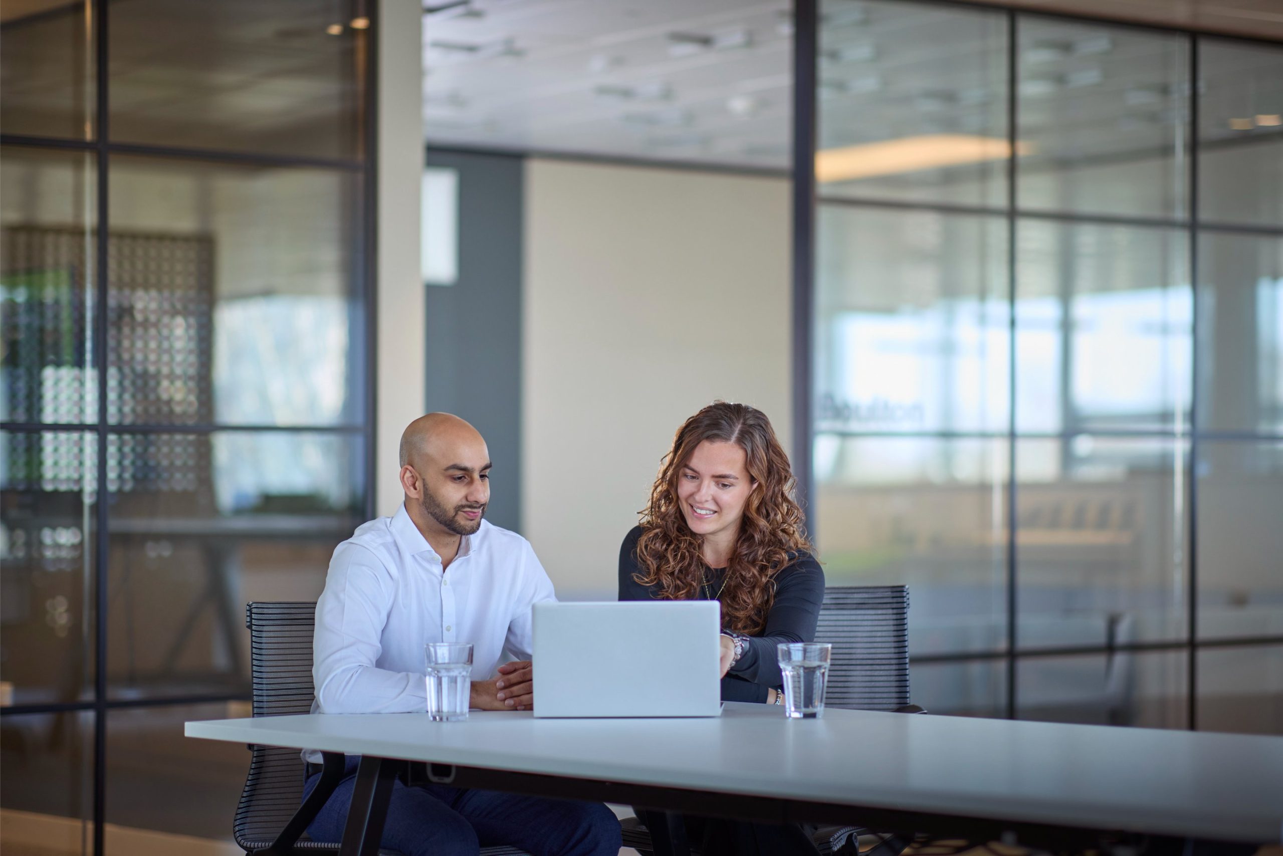 Two colleagues, a man and a woman, are seated at a table in a modern office, collaborating over a laptop, with glasses of water in front of them.