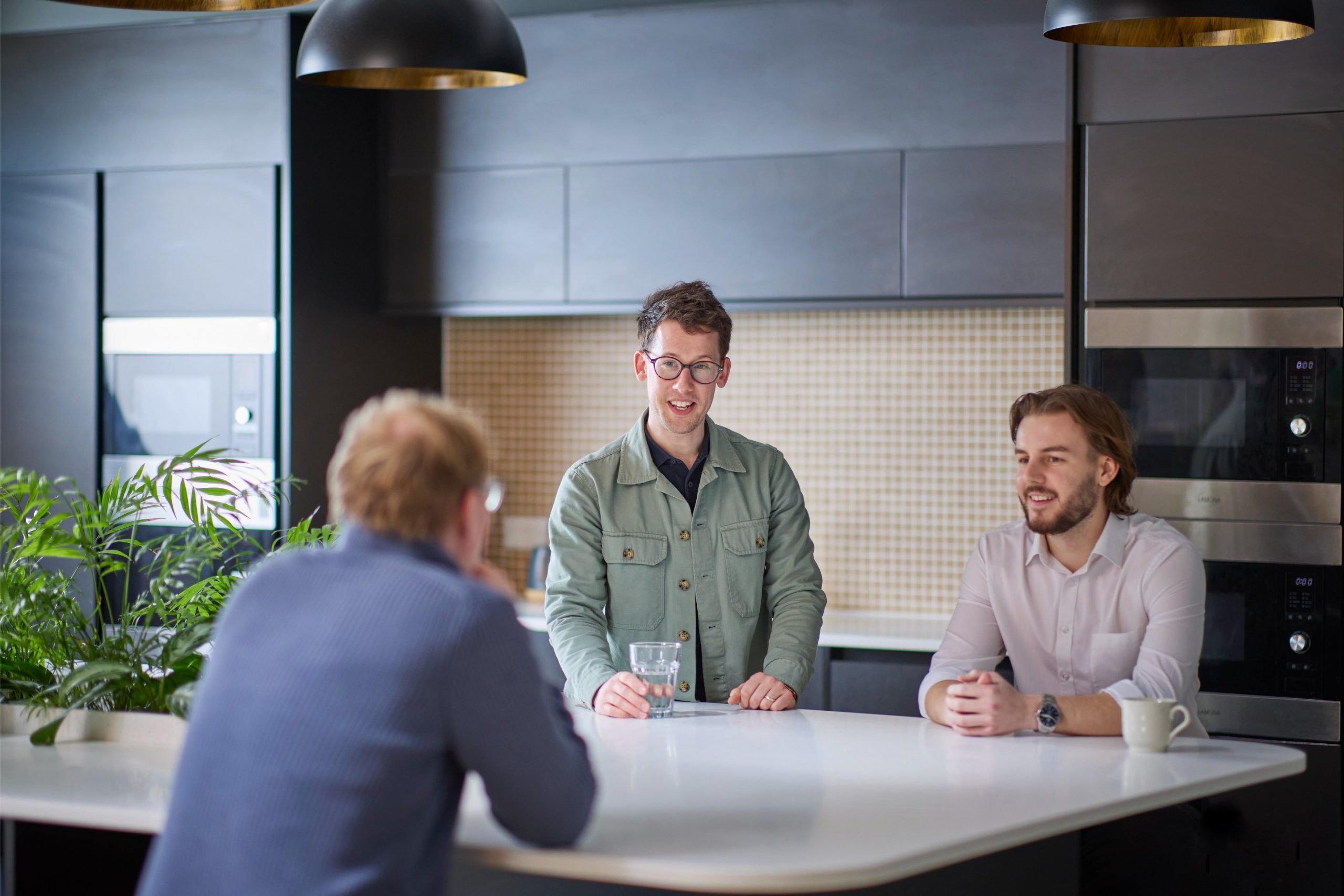 Three men engage in conversation at a modern kitchen island, with one holding a glass of water and the others seated, surrounded by plants and contemporary appl