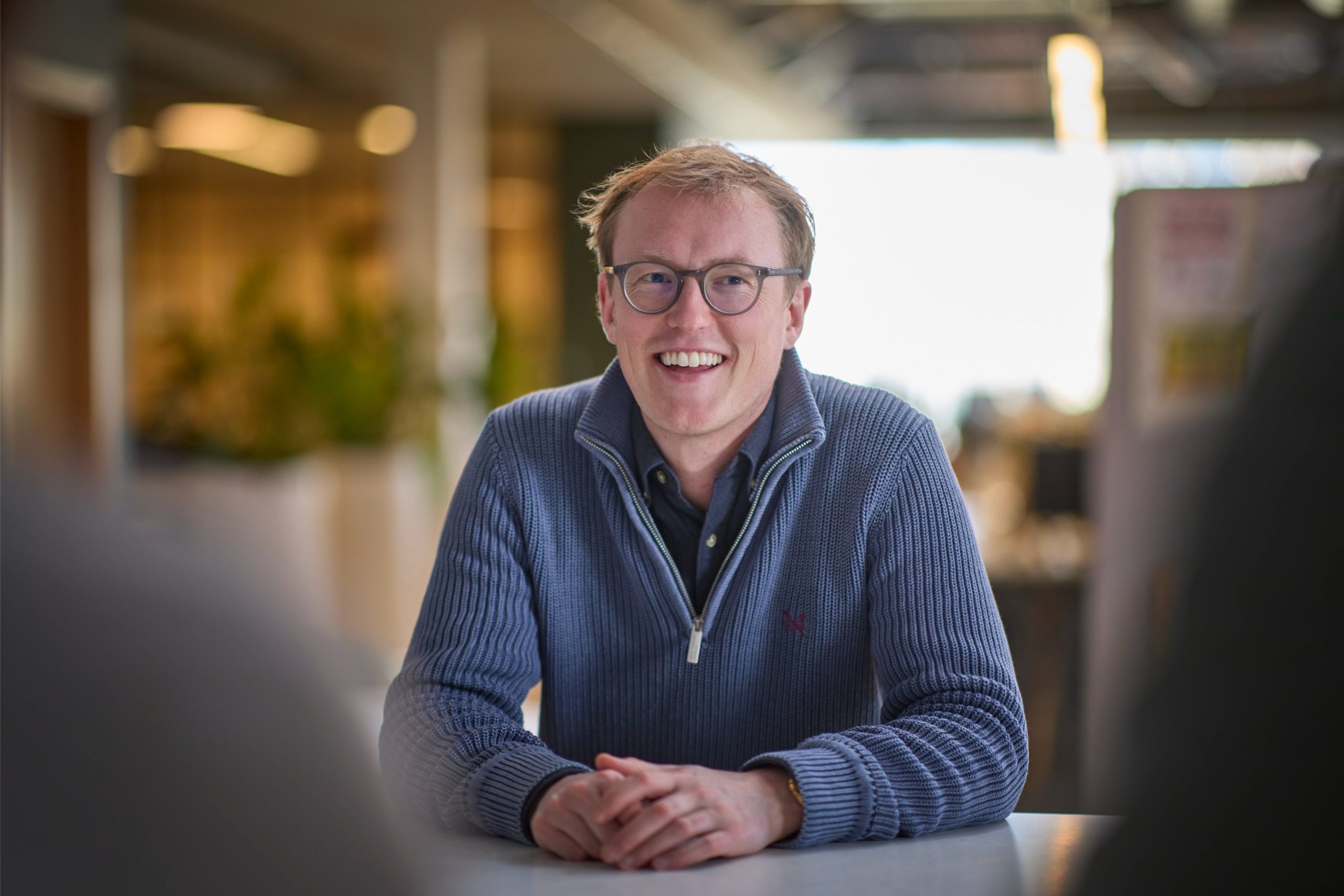 A smiling man with glasses sits at a table, wearing a blue zip-up sweater, in a well-lit office environment.