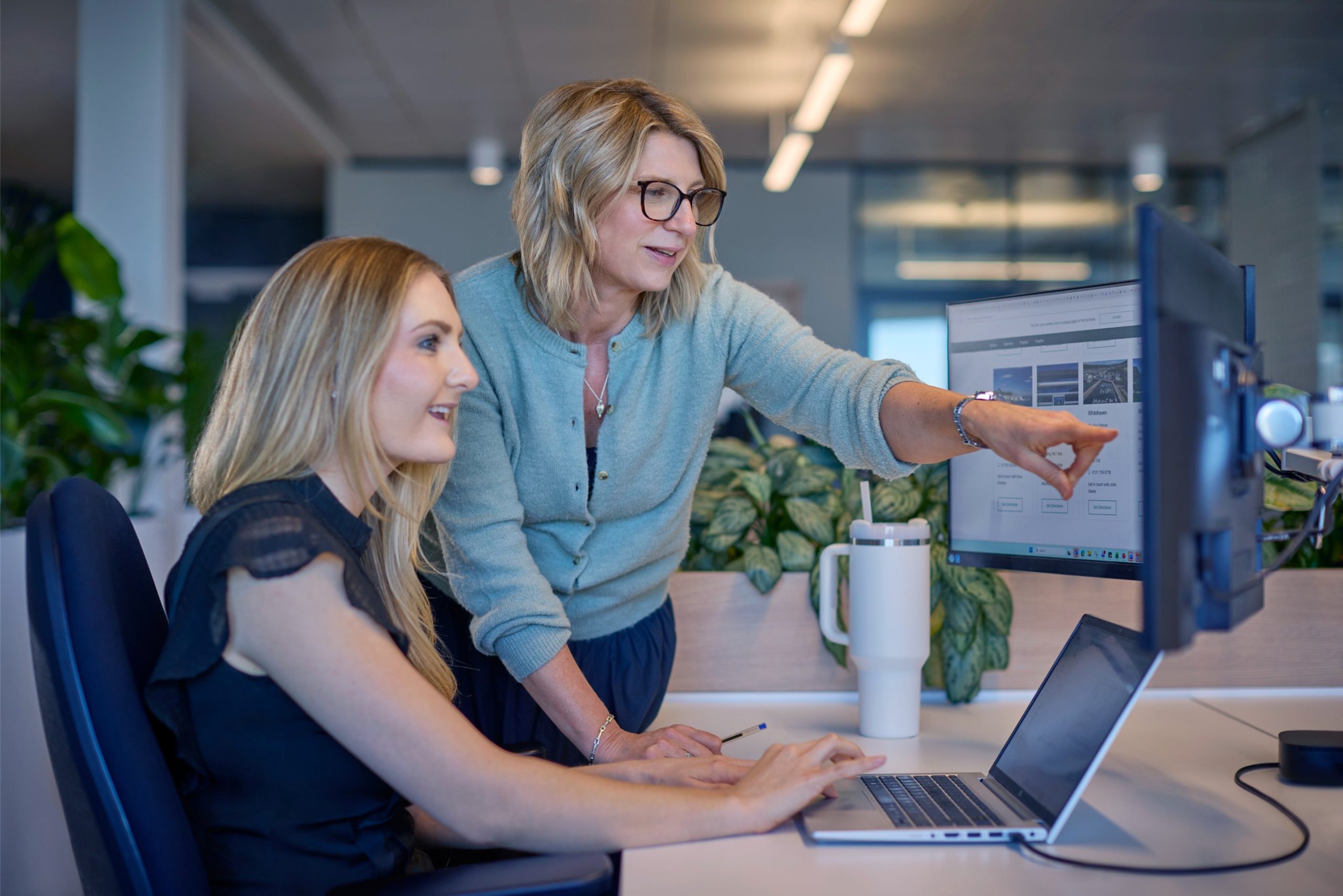 Two women collaborate at a desk, one pointing at a computer screen while the other types on a laptop, surrounded by plants and office decor.
