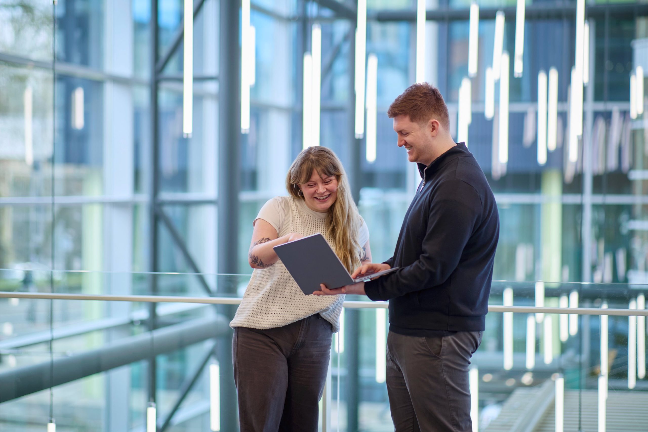 Two individuals are engaged in a discussion while looking at a laptop in a modern, well-lit space with hanging light fixtures.